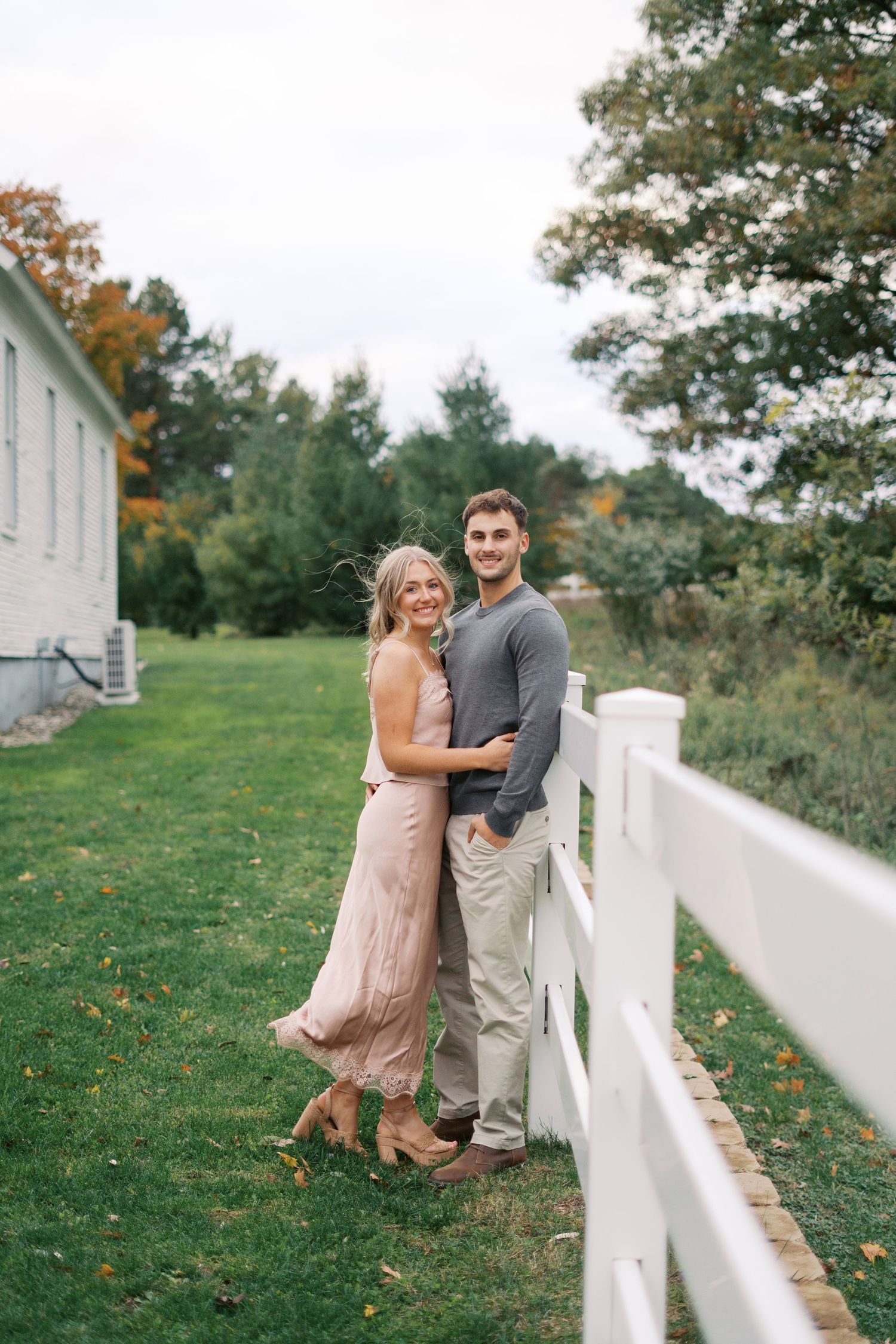 Couple poses along white fence with autumn foliage in background.