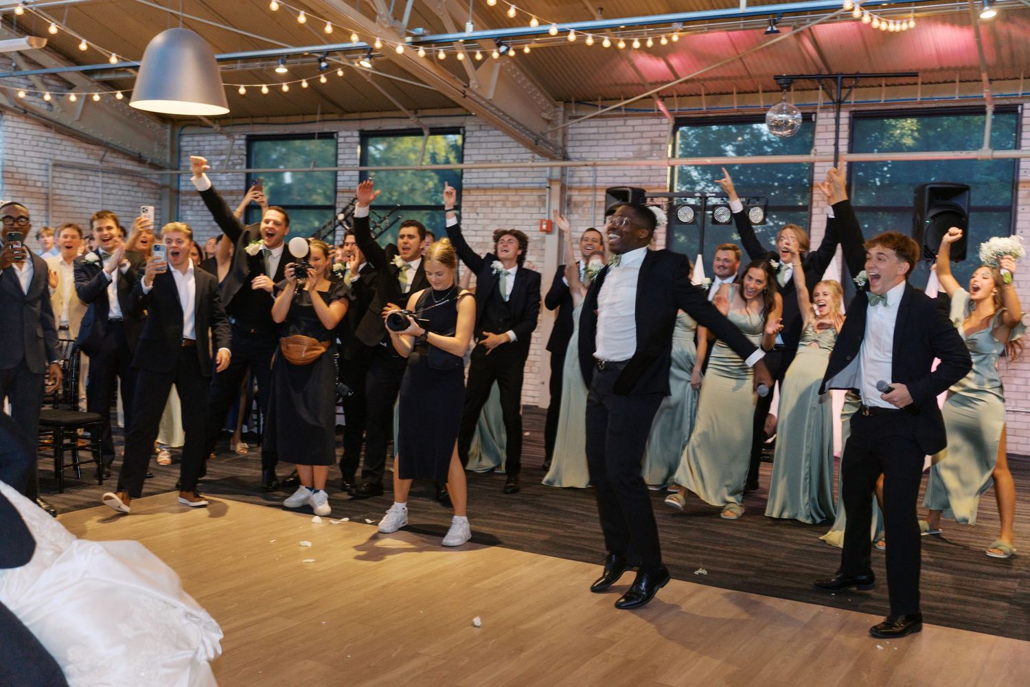 Guests dance and celebrate at a wedding reception in an industrial-style venue with string lights.