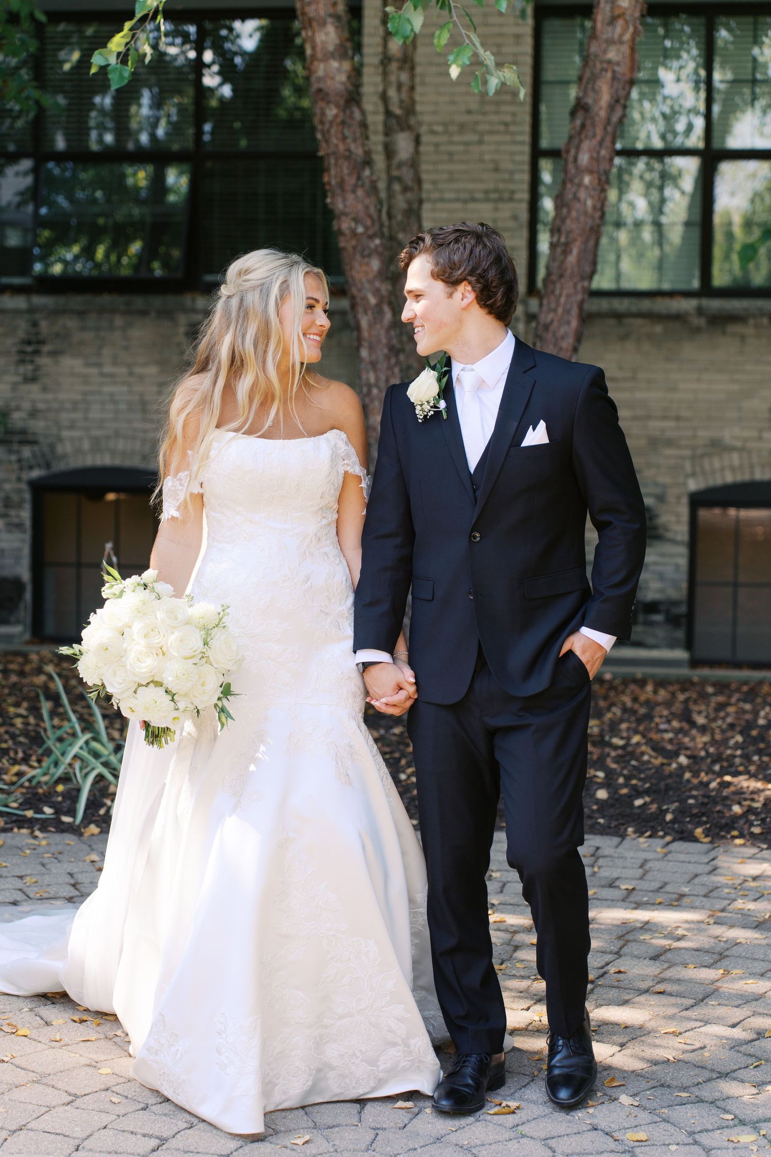 Elegant wedding couple poses on brick pathway with bride holding white floral bouquet.