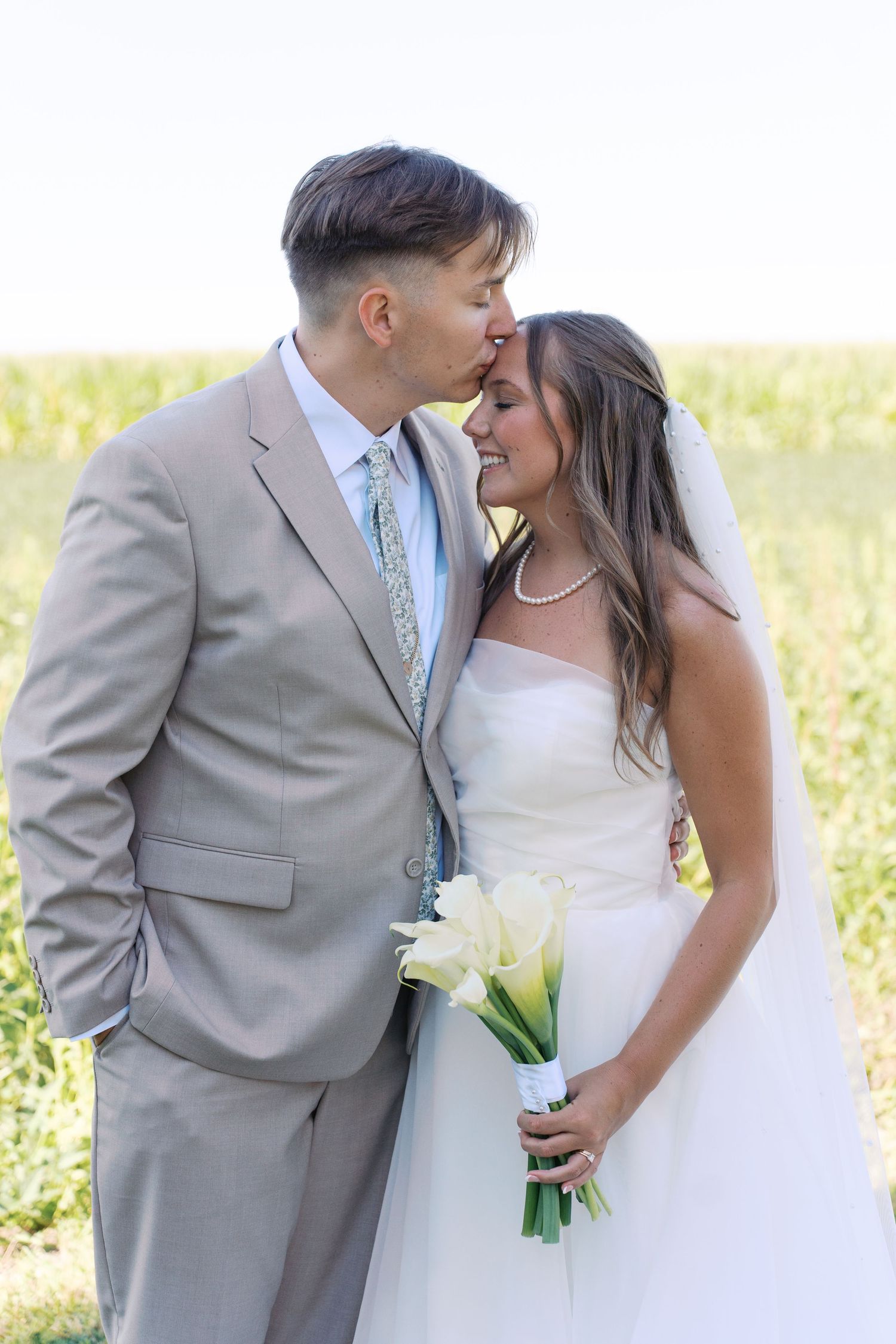 A romantic wedding couple shares intimate moments in a field, with the bride holding a white bouquet.