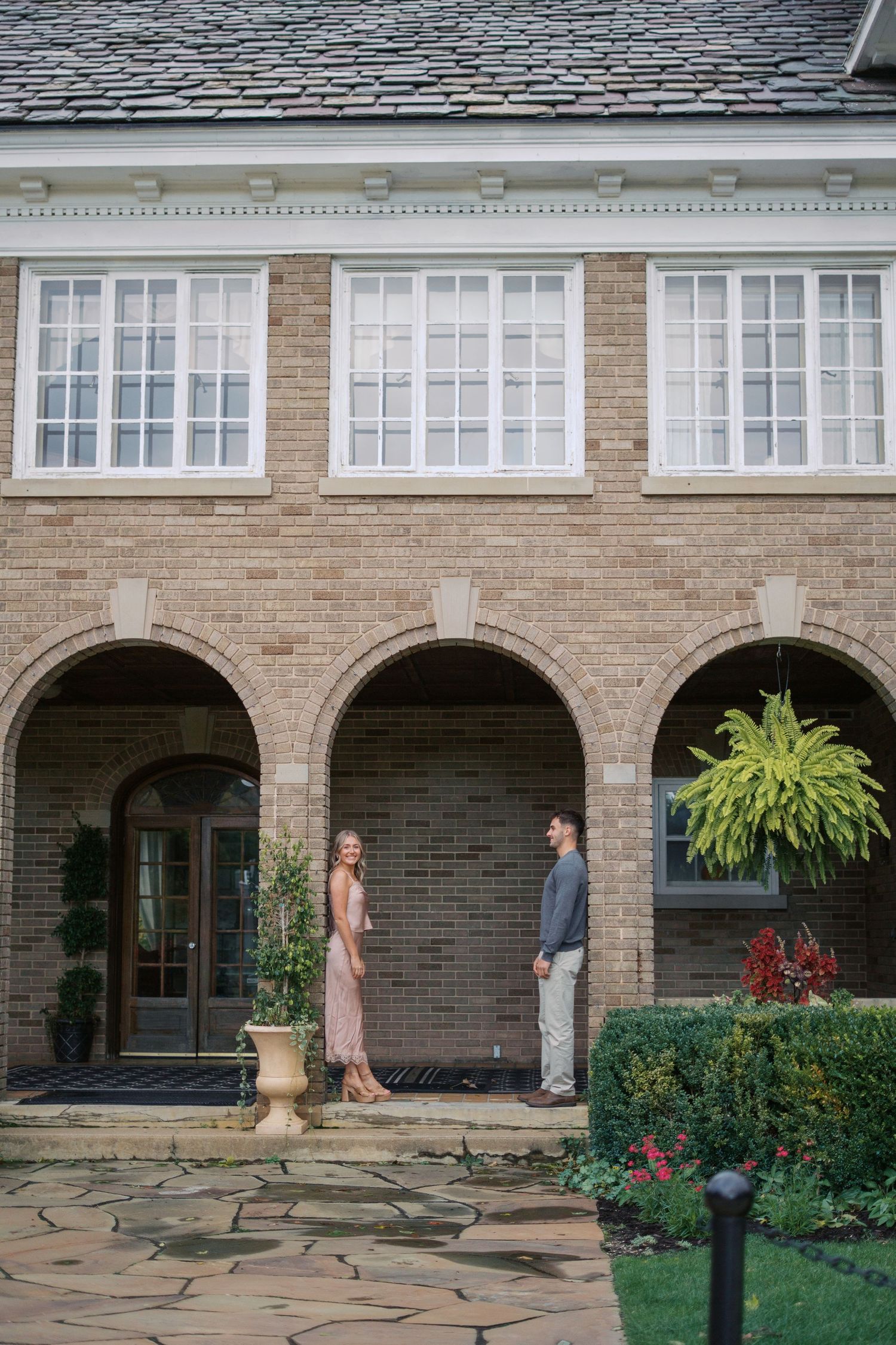 A couple standing beneath brick arches of an elegant stone building with manicured gardens.