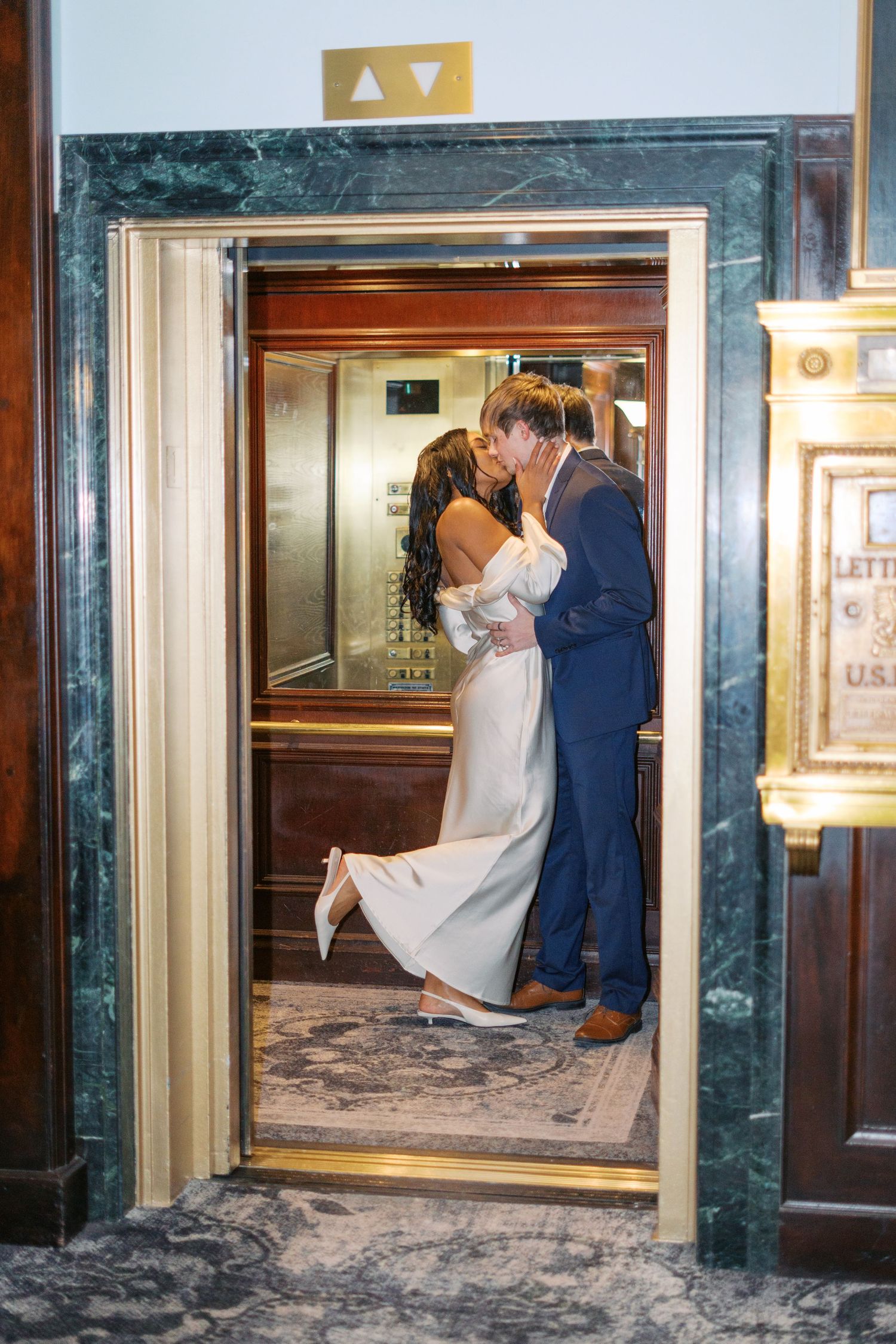 A couple embraces near gold-trimmed elevator doors in an elegant building.