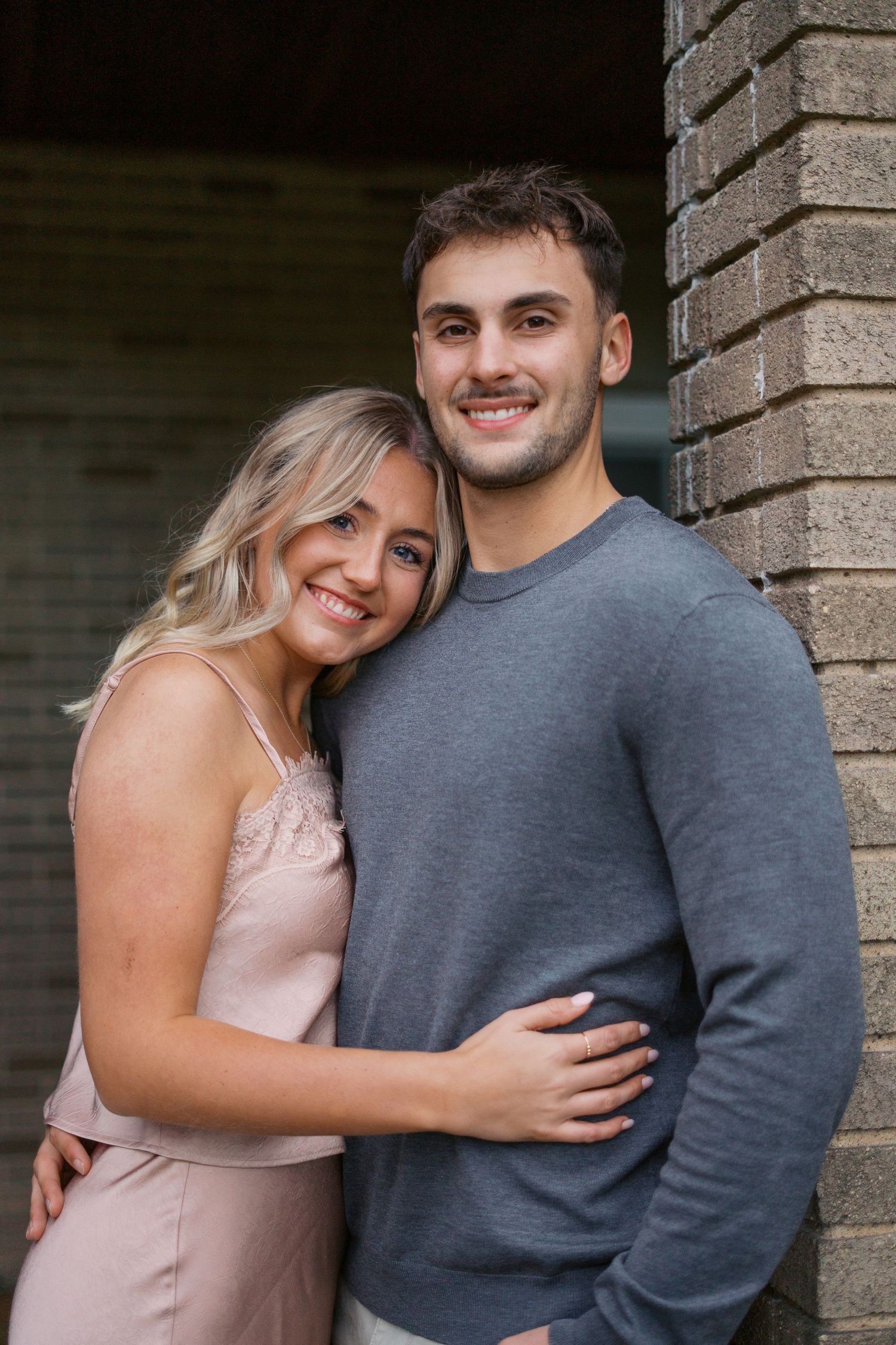 A couple embraces and smiles together against a brick wall backdrop.