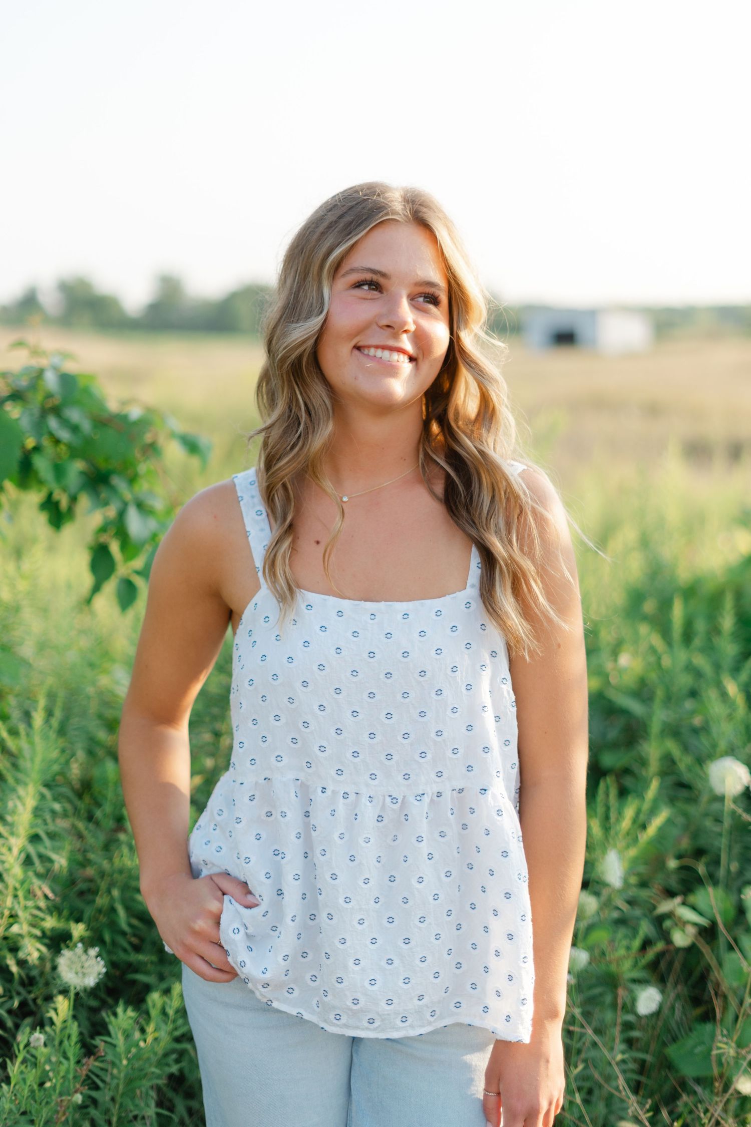 Woman in white polka dot top poses in sunny field with greenery.