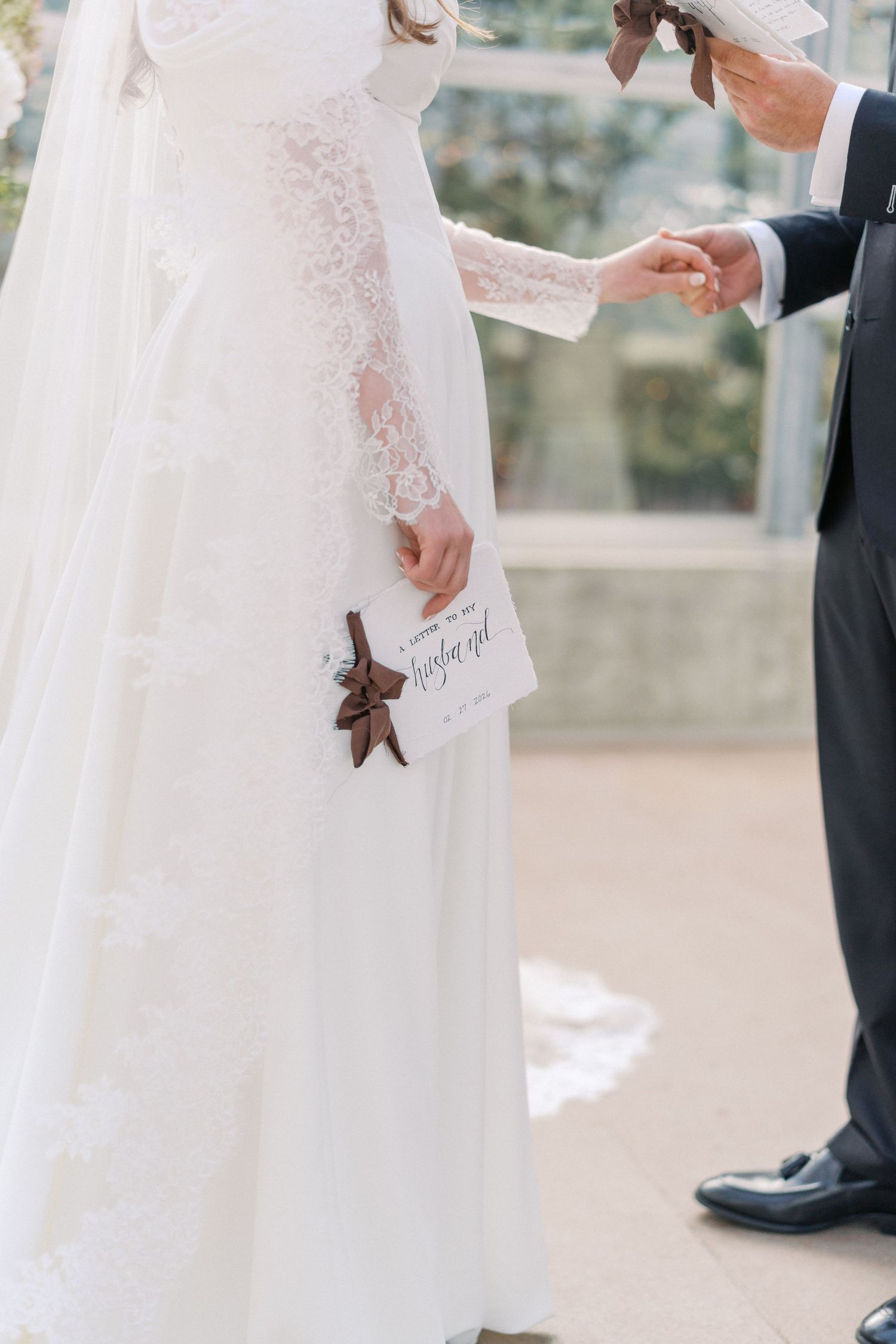 Bride in lace wedding gown holding a card and starfish while groom in black suit holds her hand outdoors.