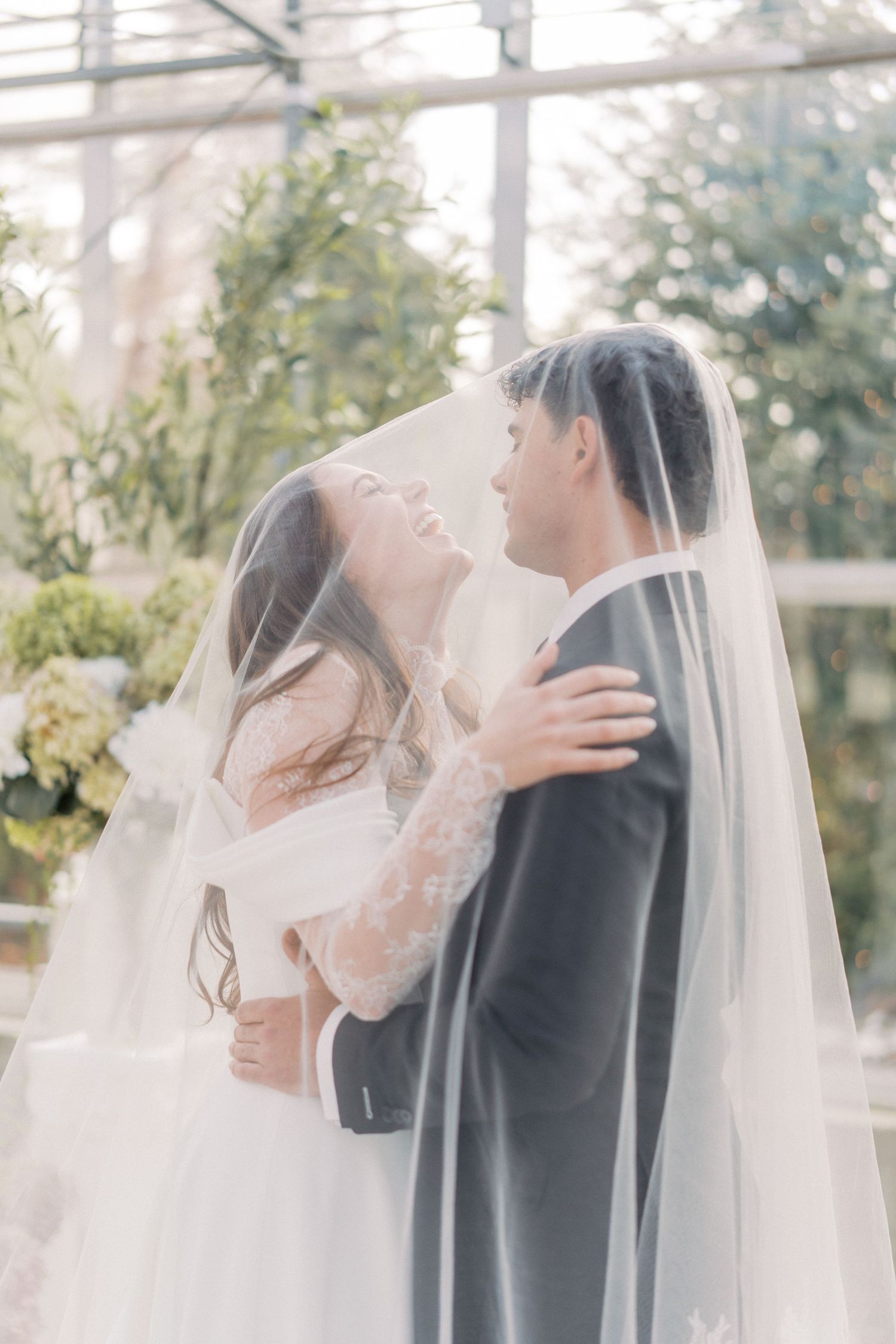 Bride and groom share romantic moment under wedding veil in a sunlit greenhouse surrounded by lush greenery.