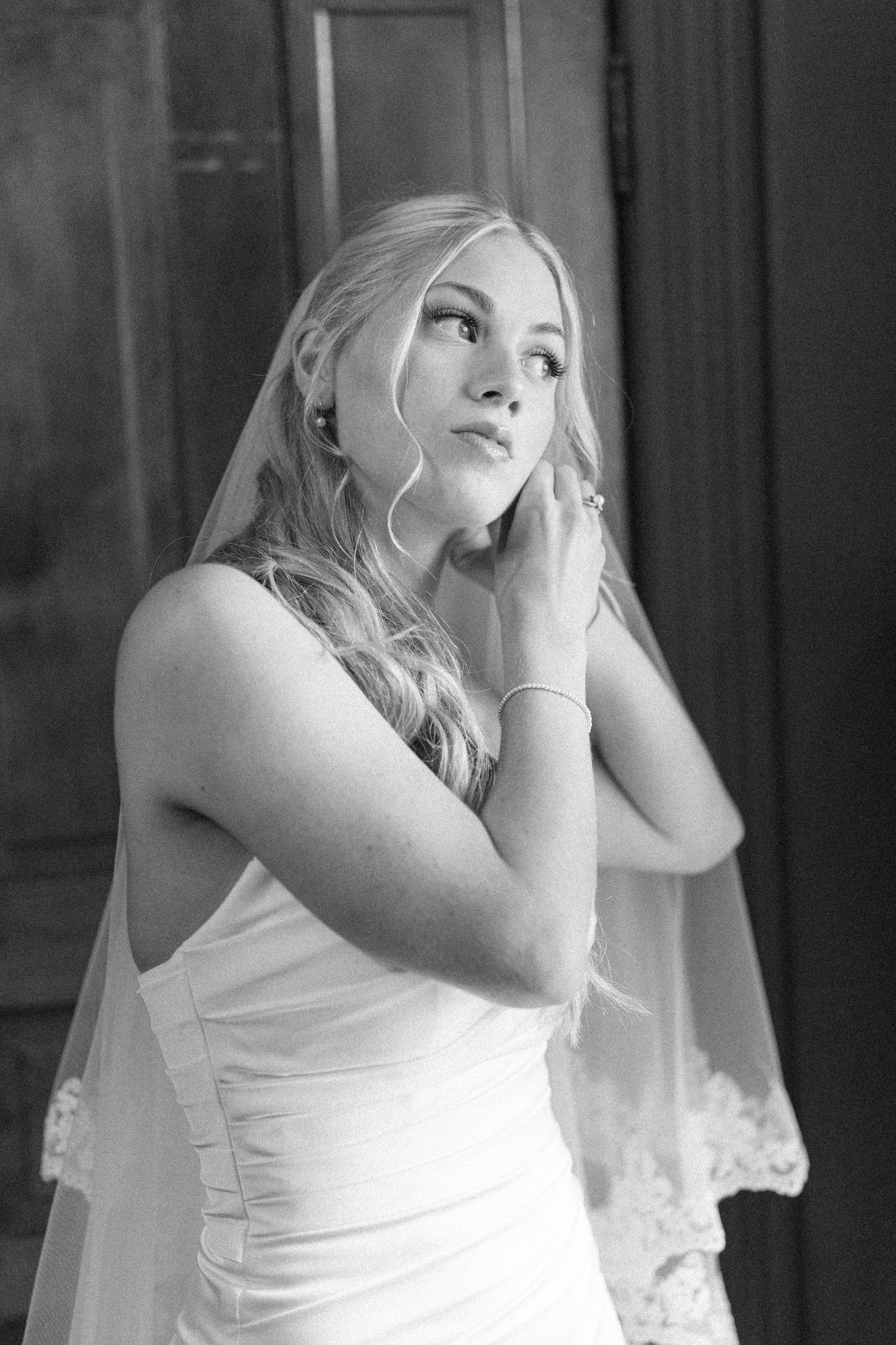 Black and white photo of a bride in a wedding dress and veil, gazing upward while adjusting her earring.