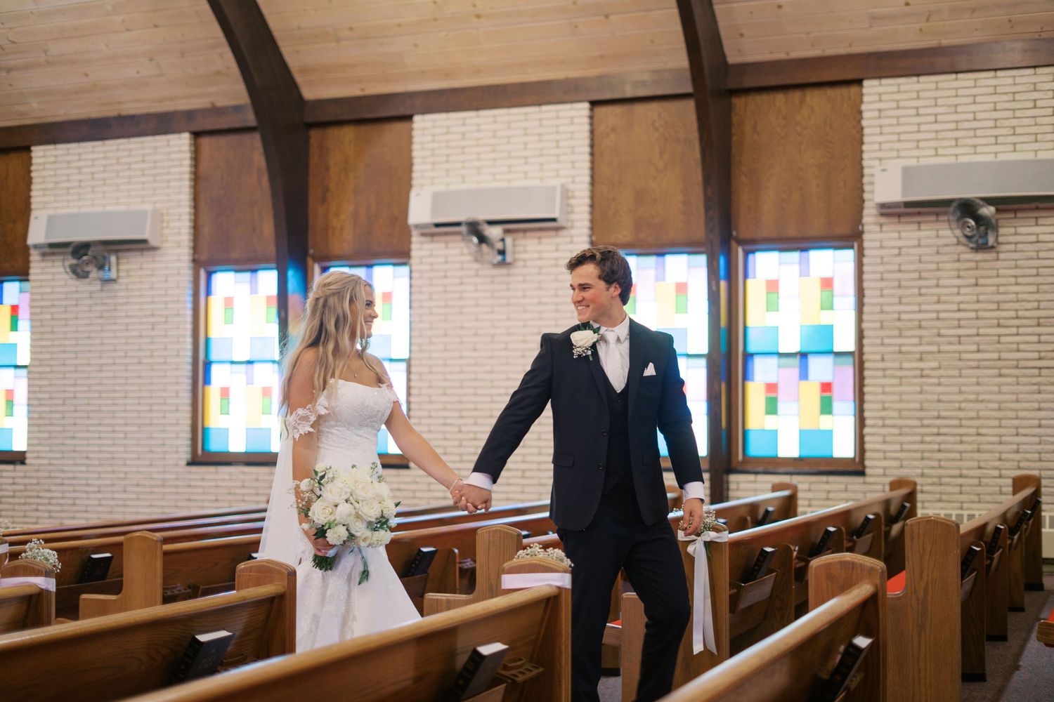 Wedding couple holding hands in church with wooden pews and stained glass windows.