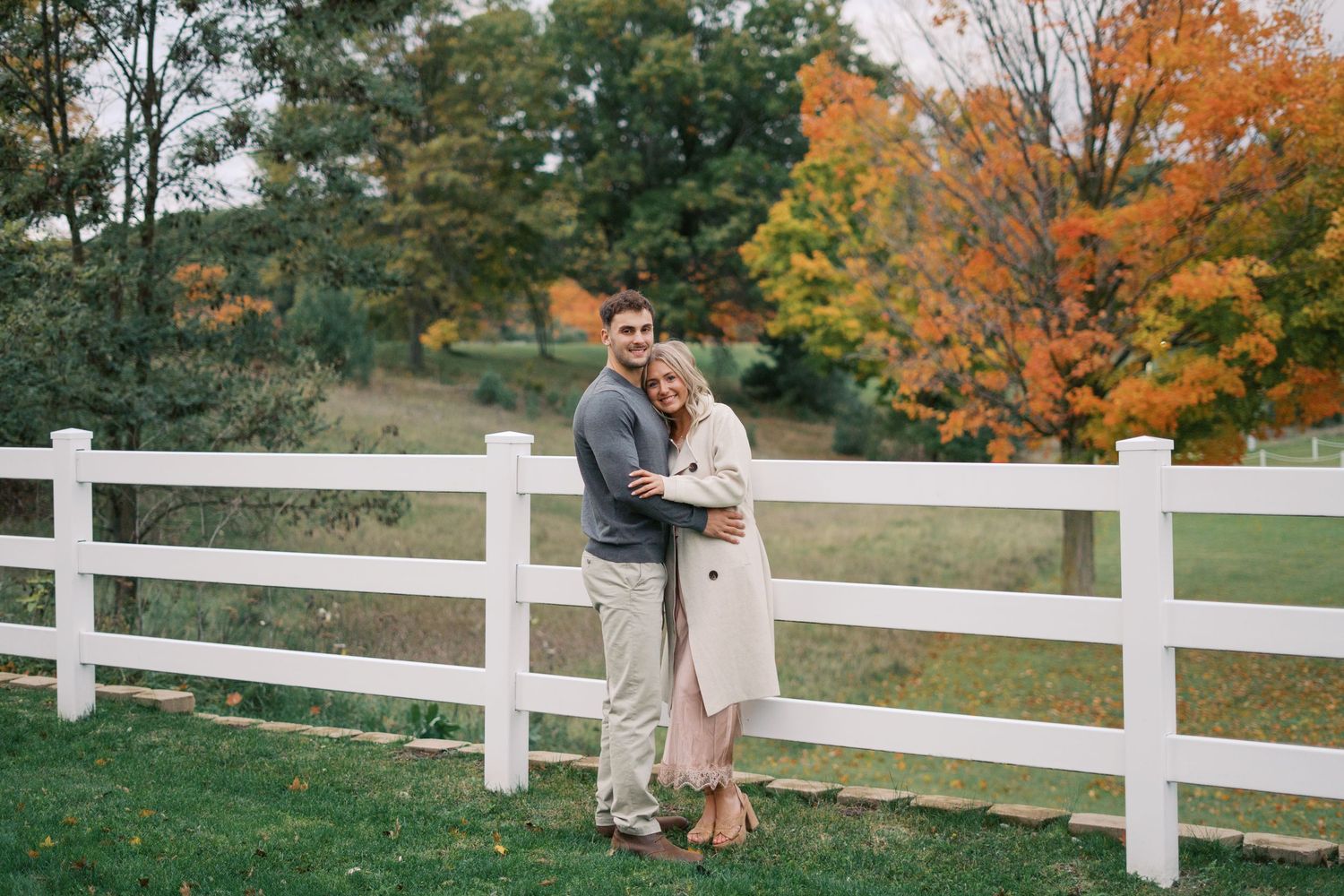 Autumn engagement photos along white fence with fall colors in background.