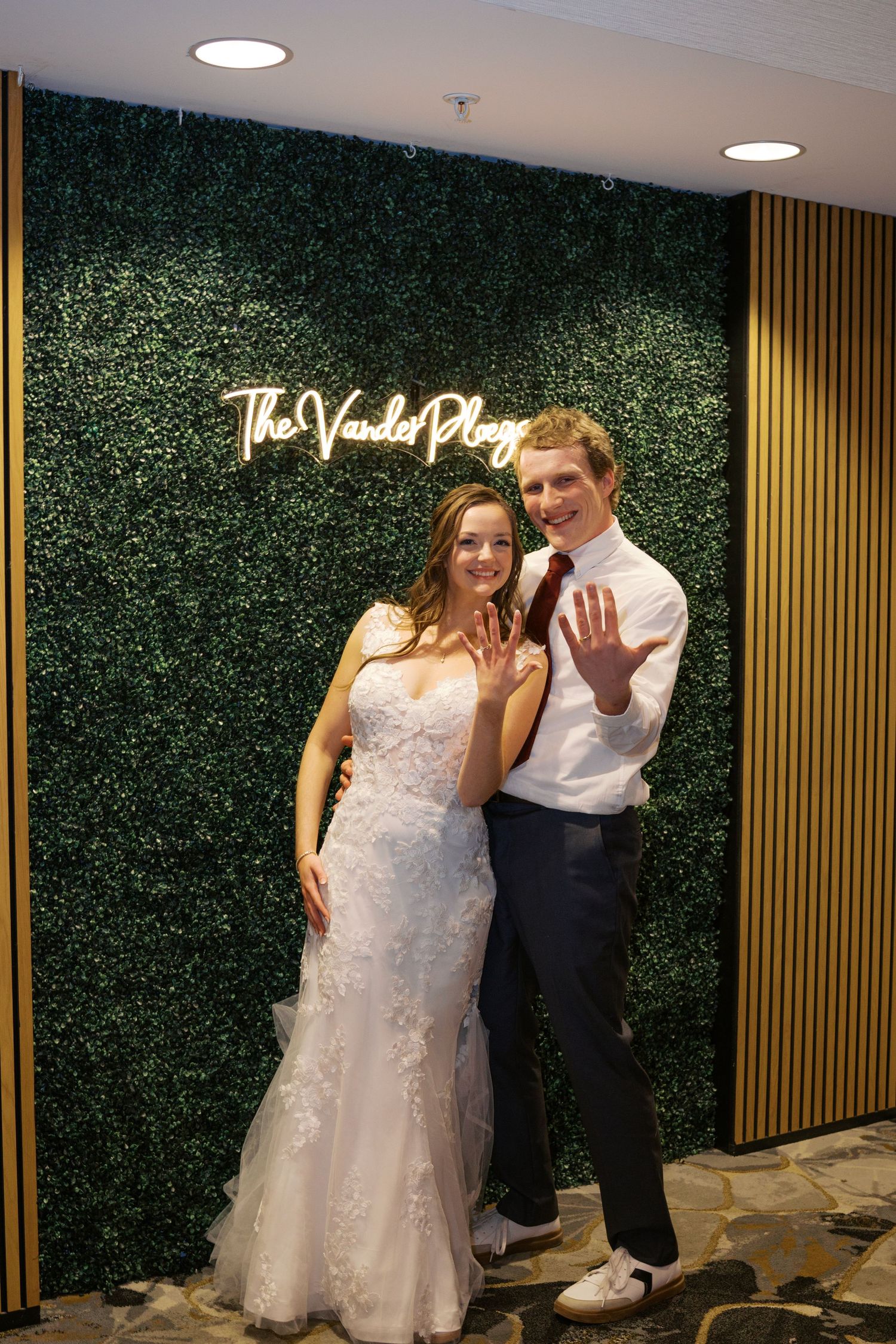 Wedding couple poses against green hedge wall with decorative lighting.