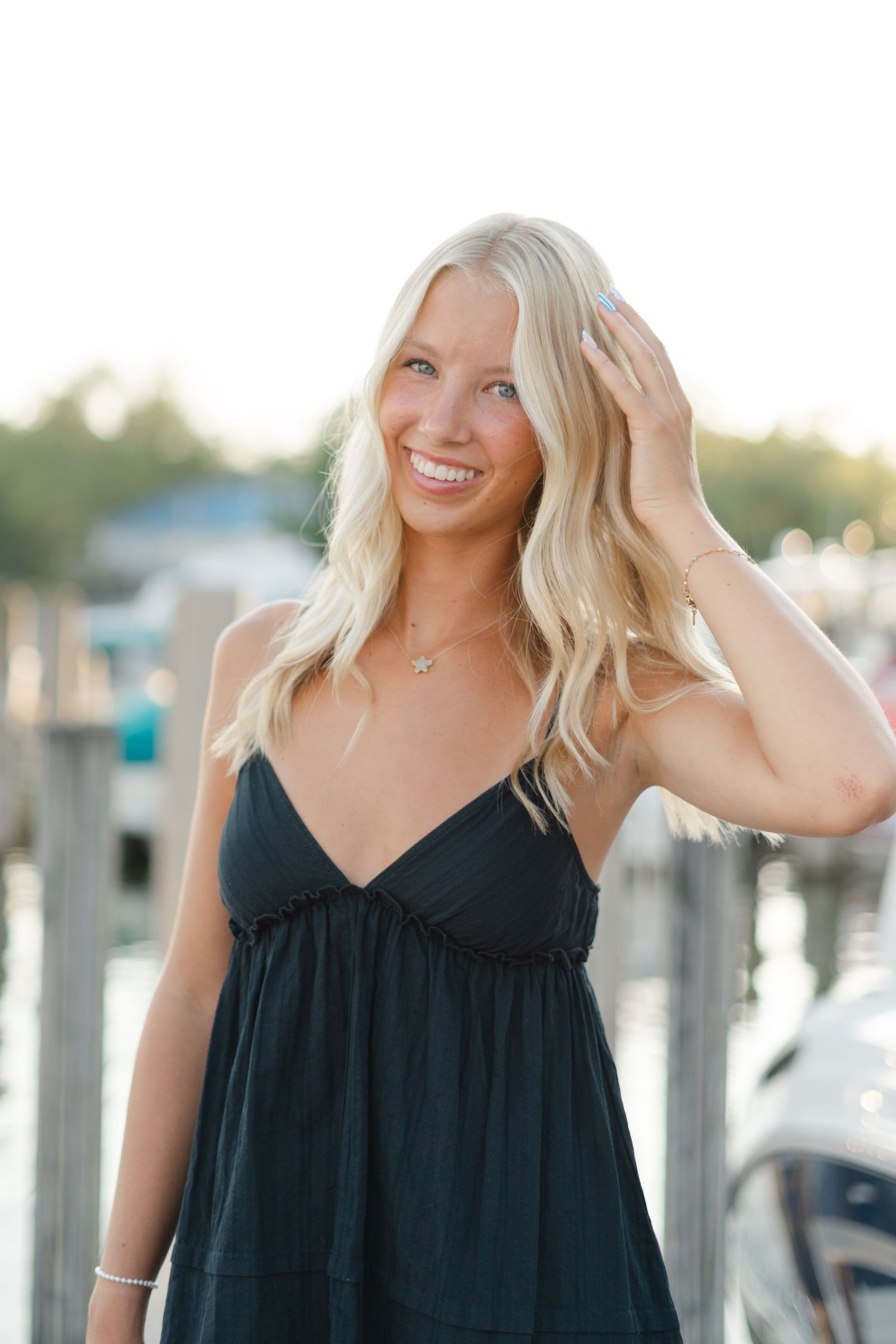 Woman in black summer dress poses casually by marina docks.