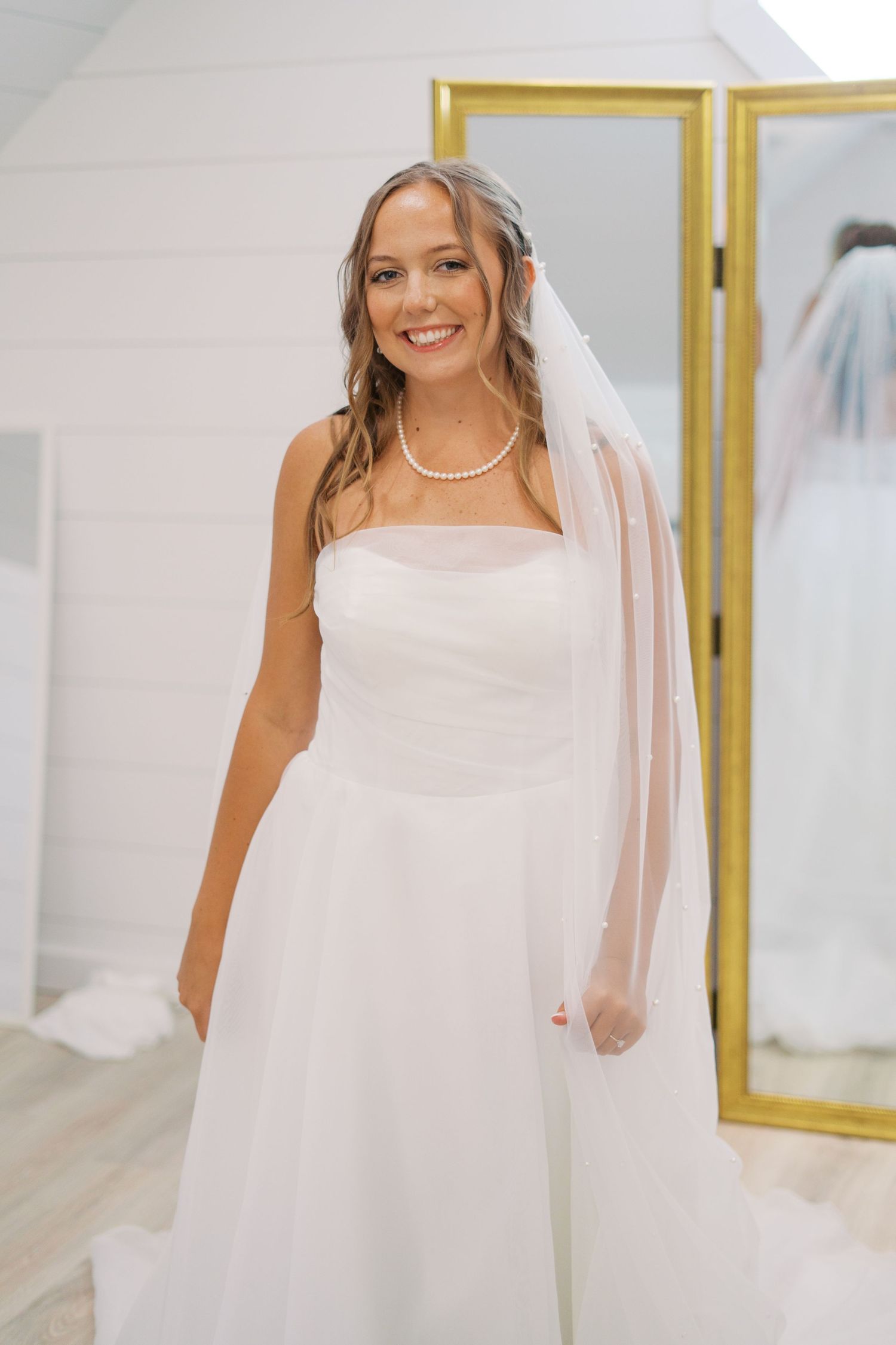 A bride in a classic white wedding gown poses with her veil in front of gold-framed mirrors.