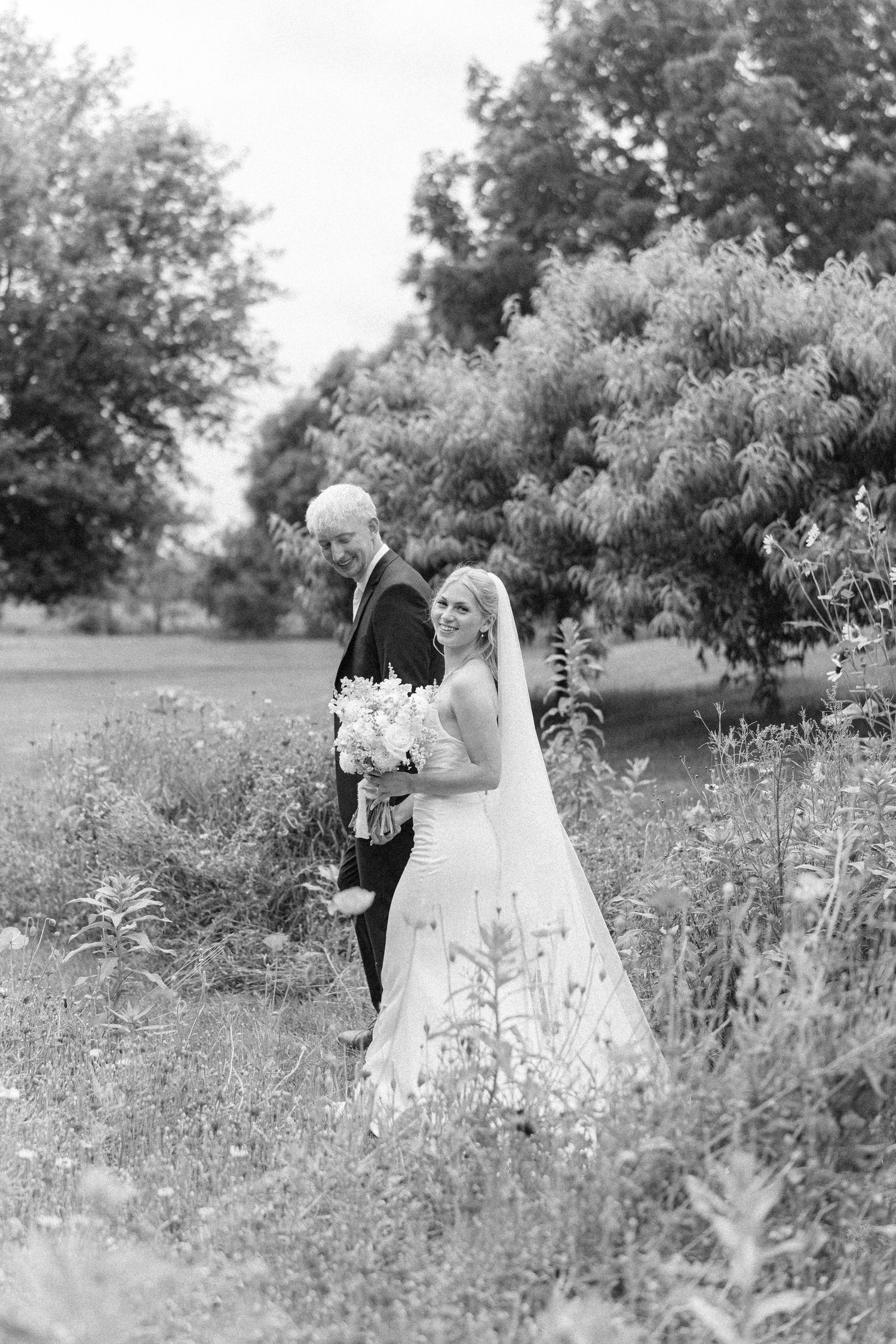 Black and white photo of a smiling bride in a veil holding a bouquet, standing beside a groom in a lush outdoor setting.