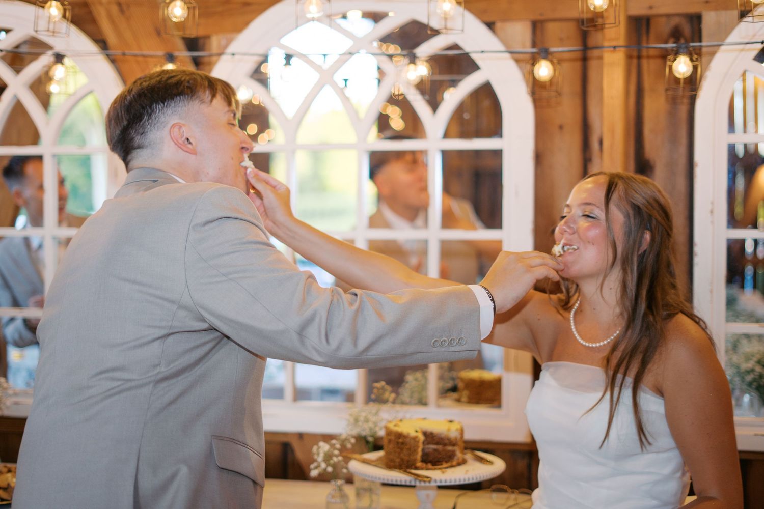 Playful cake feeding moment between newlyweds in rustic barn reception venue.