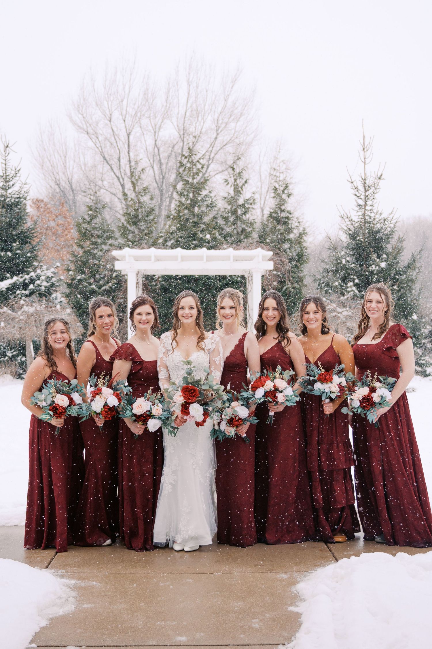 Winter wedding party photo with burgundy dresses against snowy evergreen backdrop.