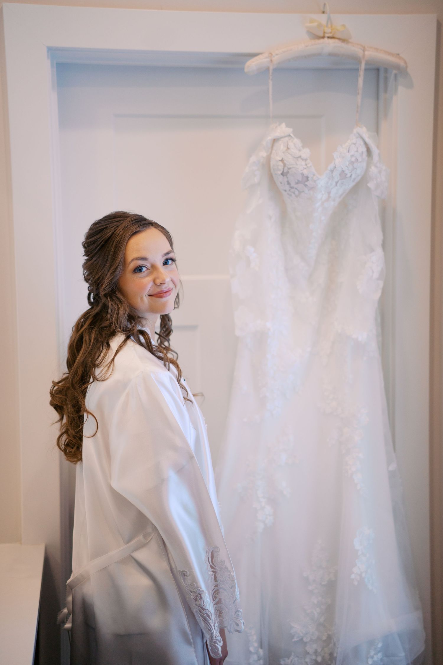 Wedding dress hangs on display while bride prepares in white robe.