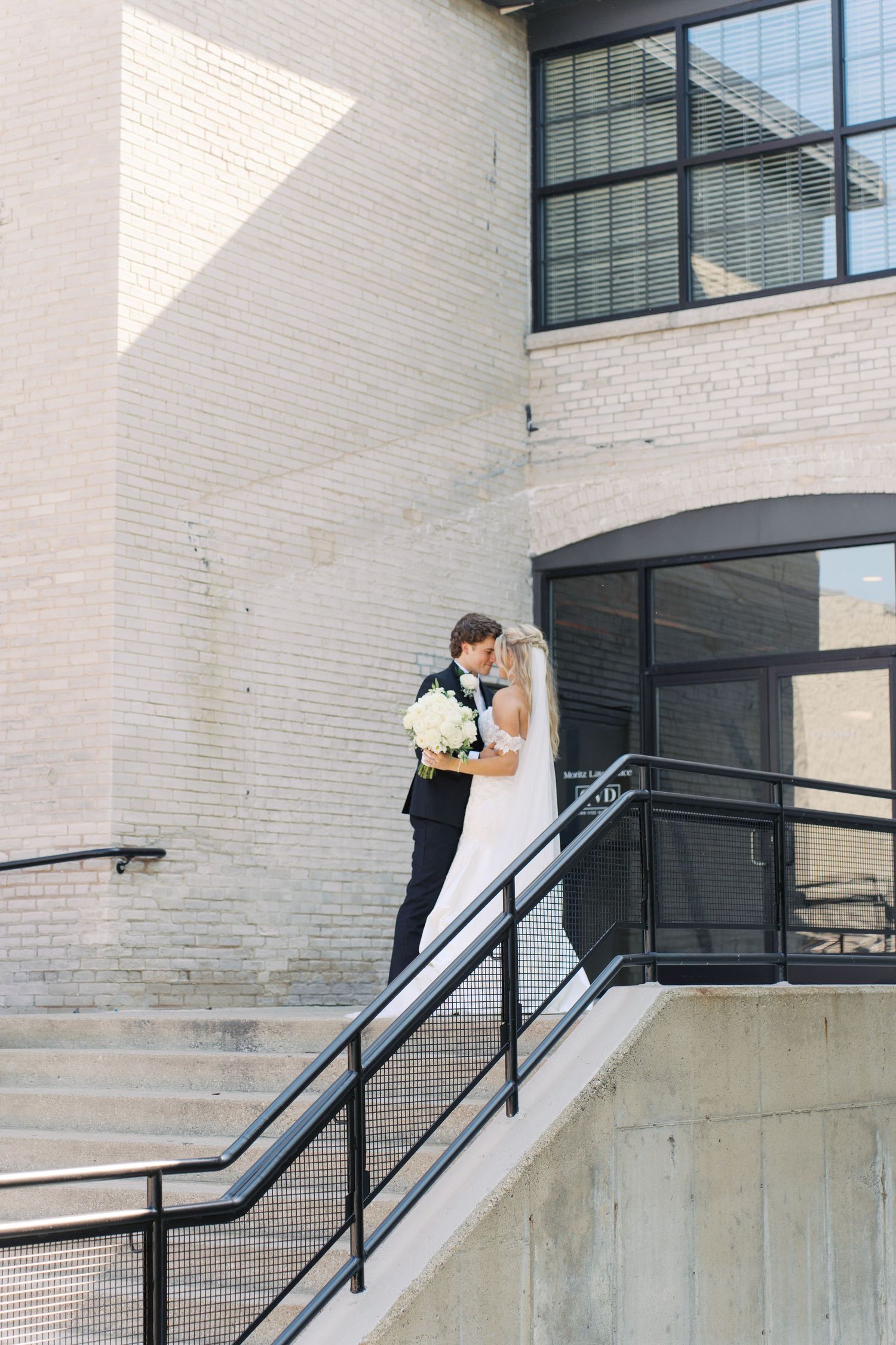 Wedding couple shares intimate moment on modern building staircase with white brick walls.