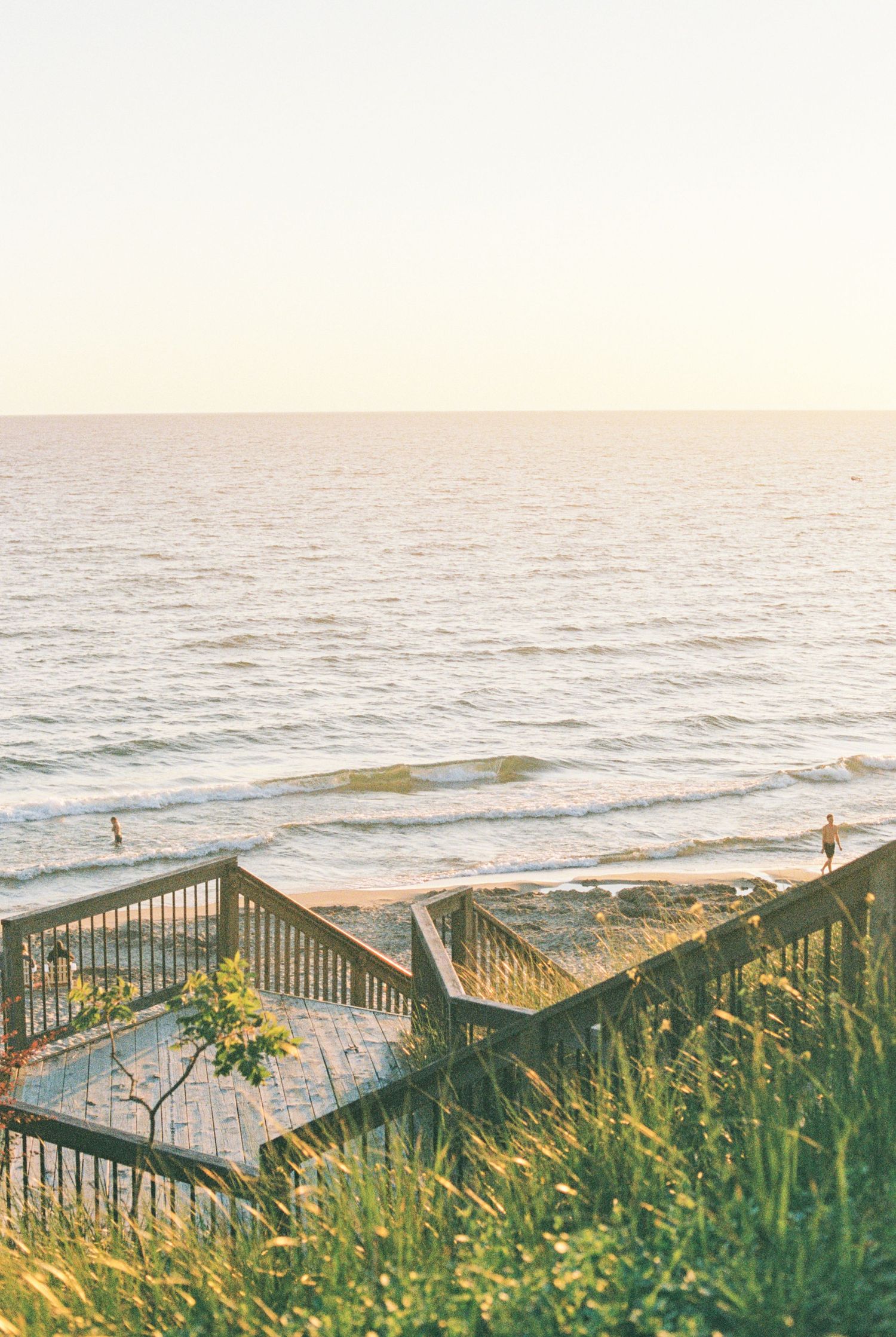 Wooden beach stairs descending to a sandy shoreline with gentle ocean waves under warm golden sunset light.