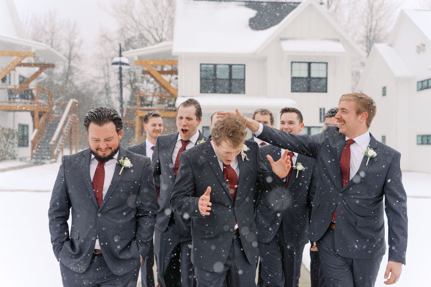 Wedding party in dark suits laughs together during snowy photo session.