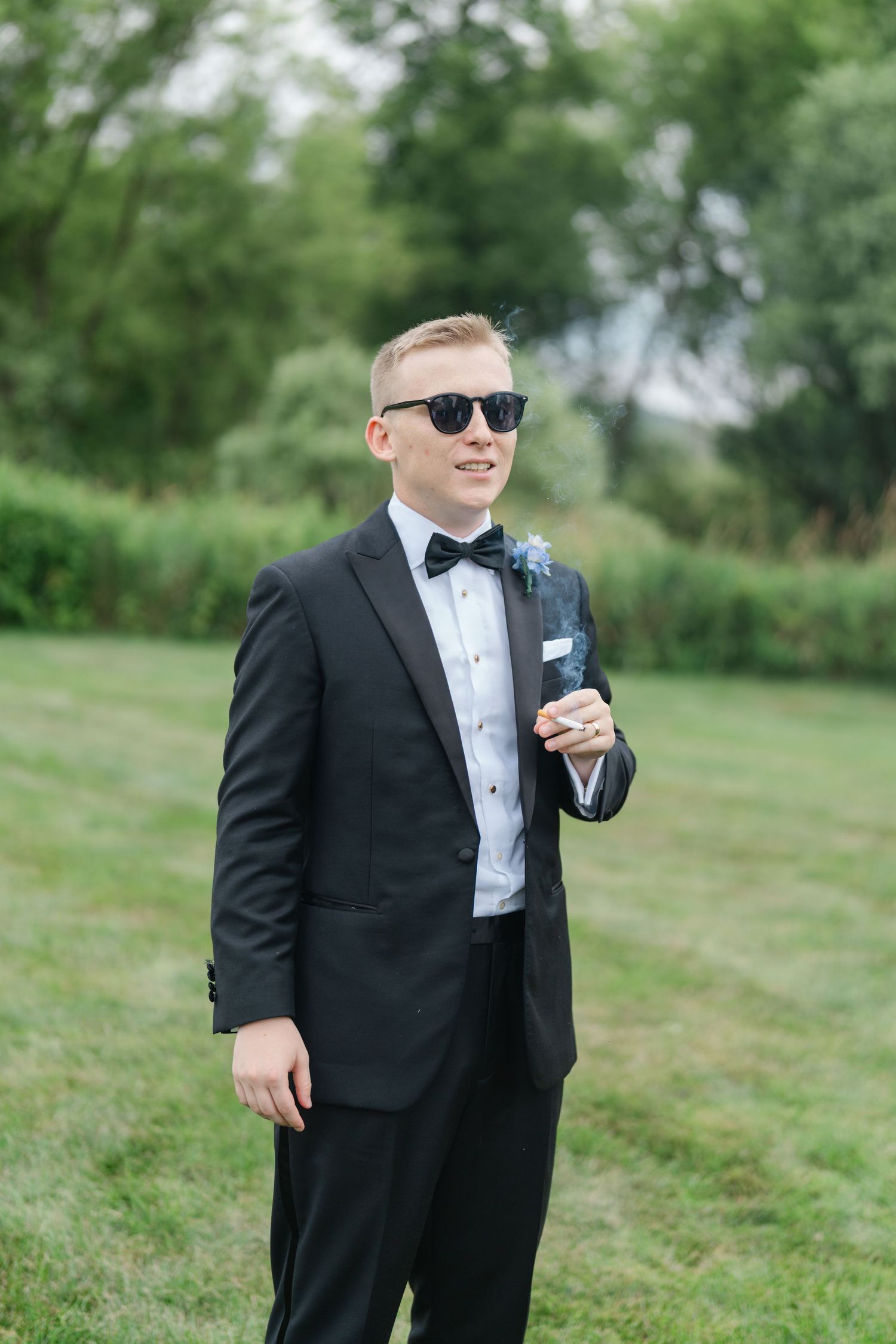 Groom in classic black tuxedo poses confidently on grassy hillside wearing sunglasses.