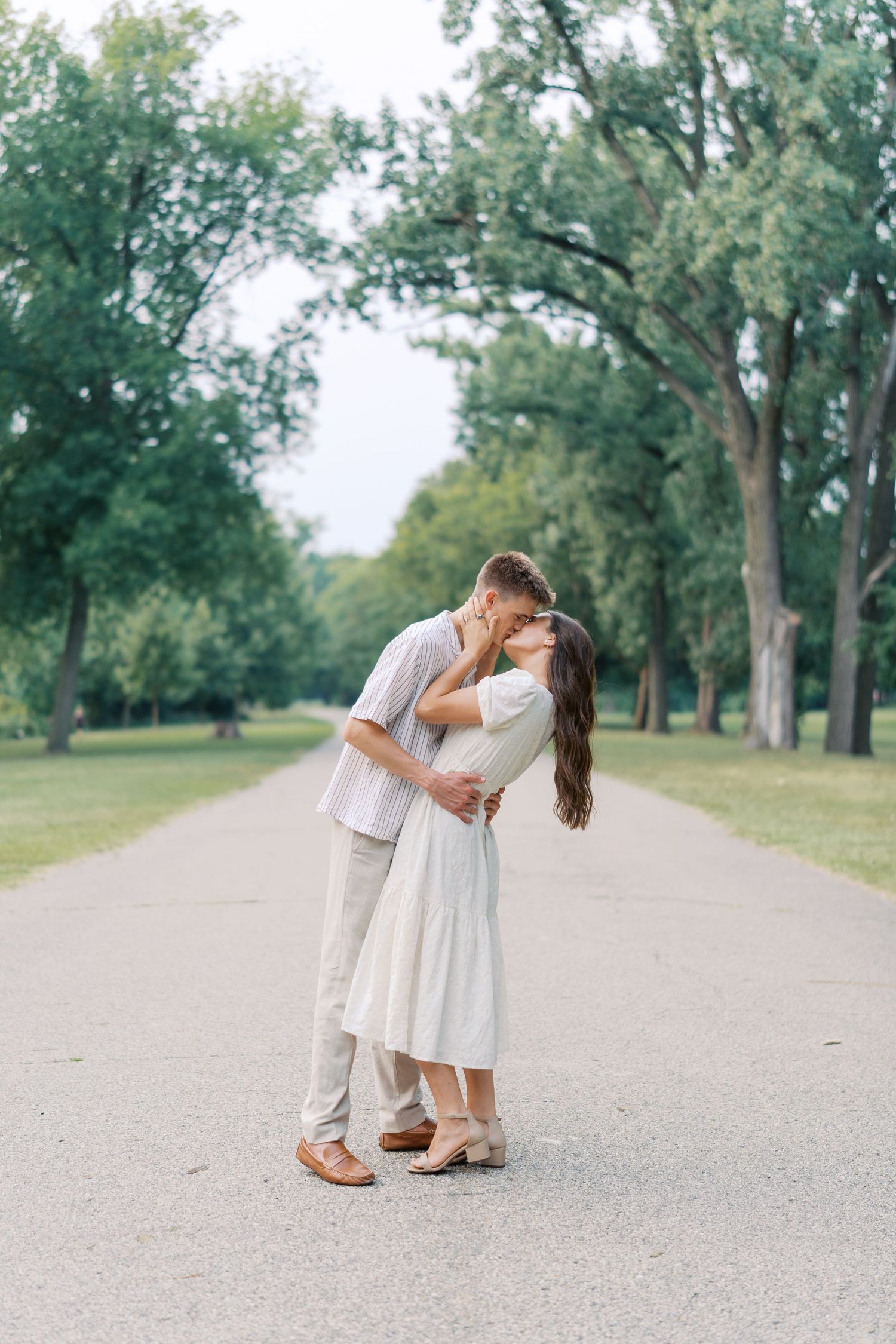 Couple sharing a romantic kiss on a tree-lined path, woman in white dress, man in light casual outfit, lush green park.