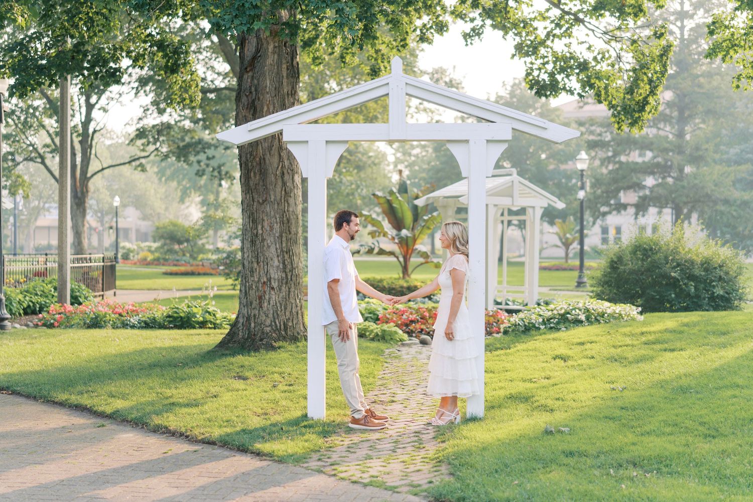 Couple holding hands under white pergola in lush garden park setting on sunny day.