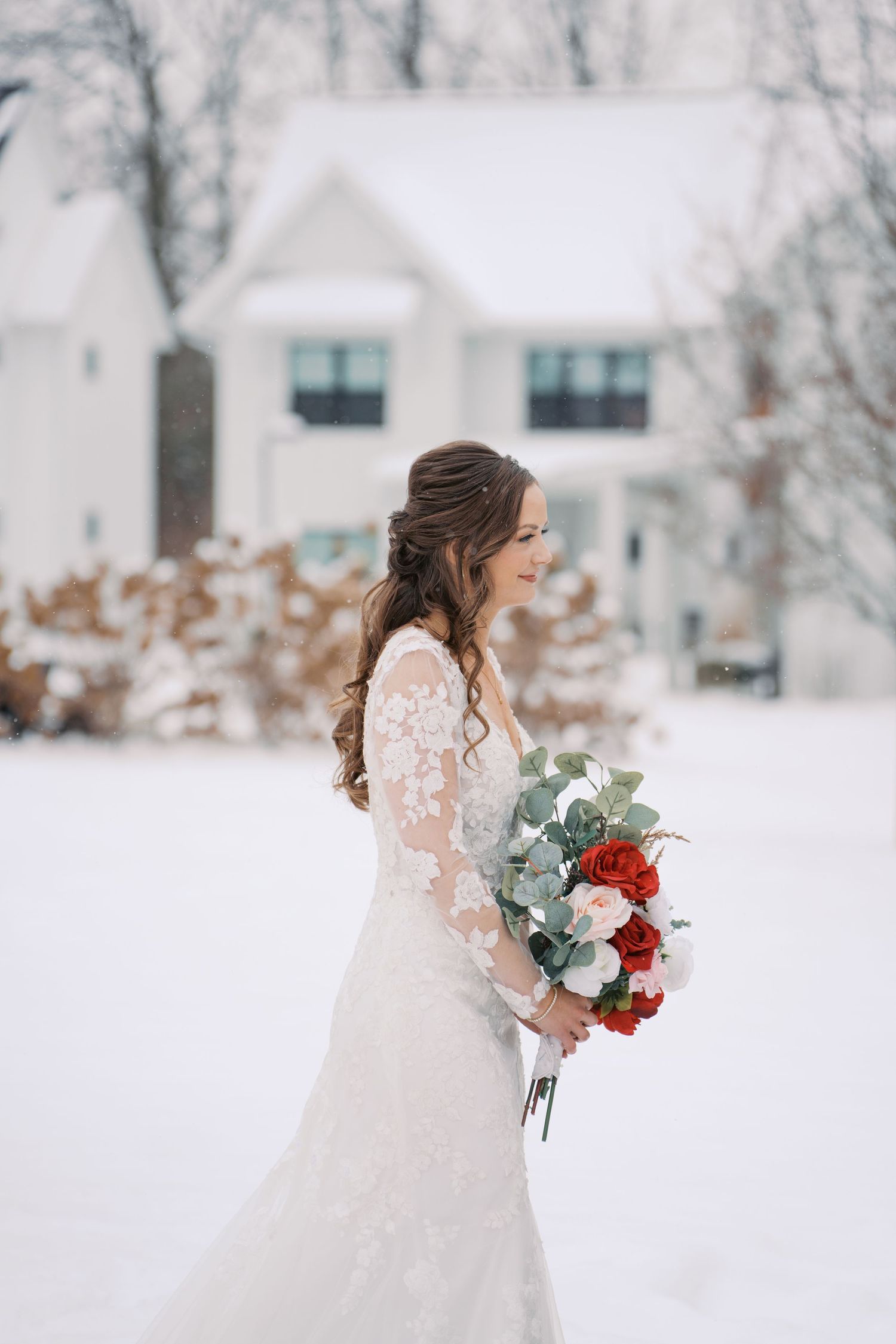 Bride in lace gown holds red and white bouquet against snowy winter backdrop.