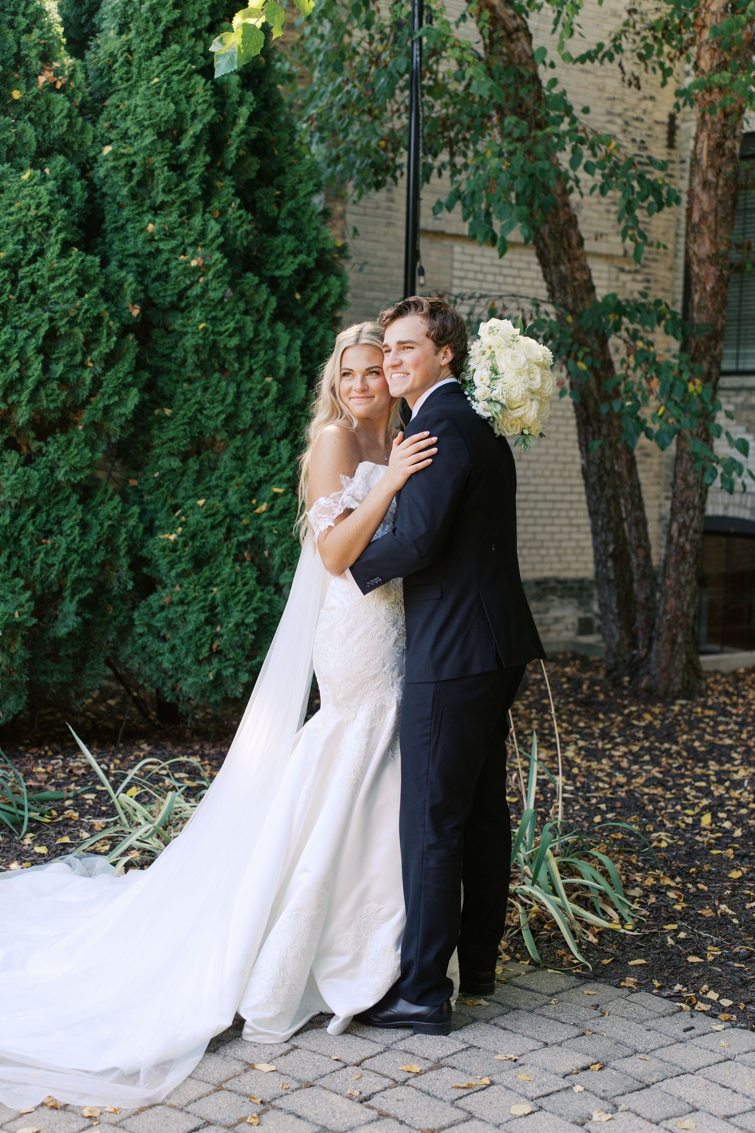 Newlyweds embrace on stone pathway surrounded by lush greenery and brick building.