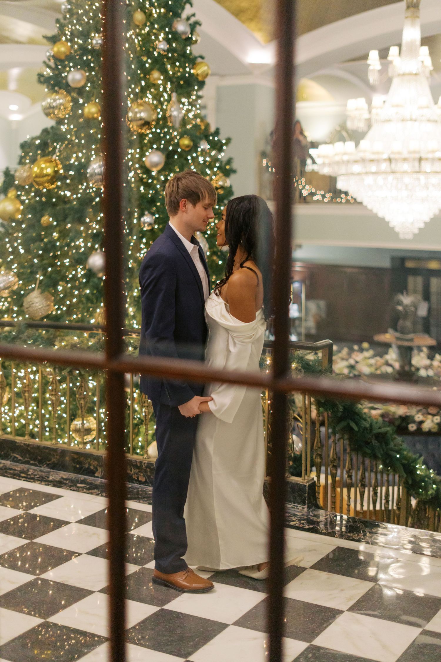 Wedding couple stands on checkered floor near decorated Christmas trees and chandeliers.