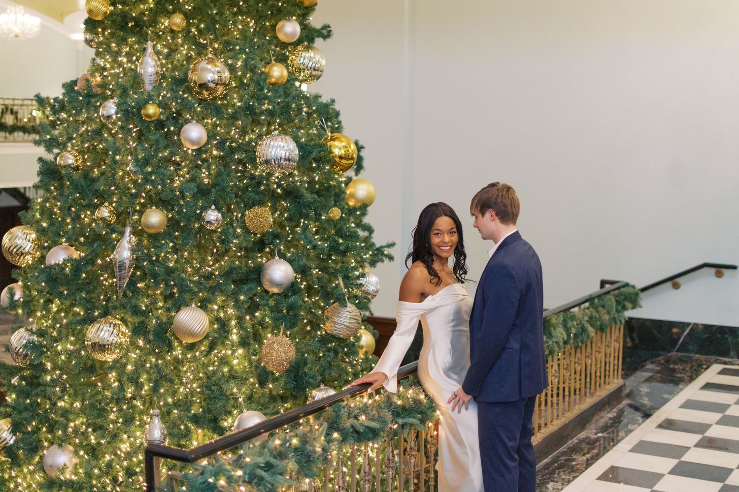 A couple stands together near a festive Christmas tree decorated with gold and silver baubles.
