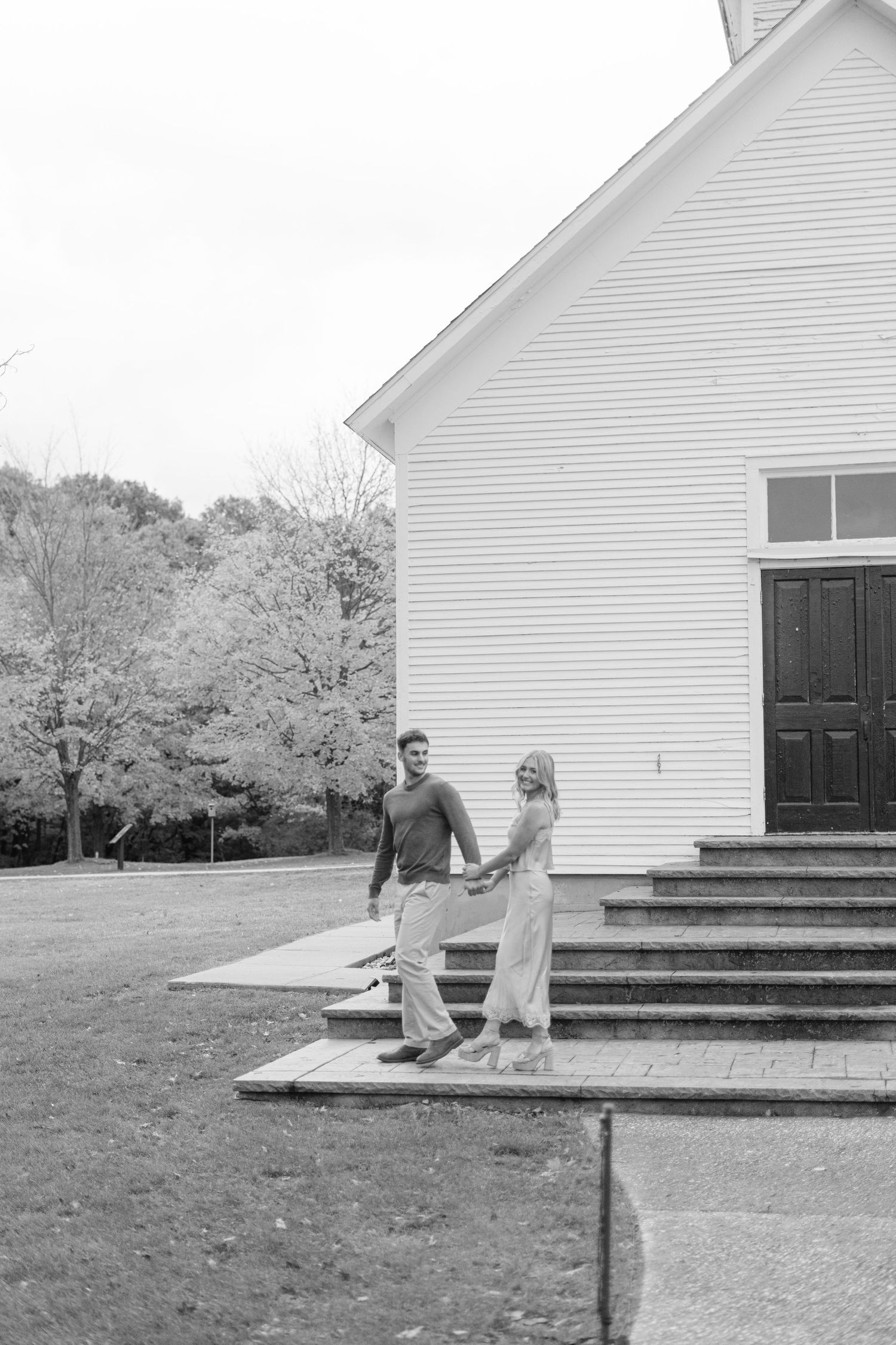 A couple shares a playful moment on the steps of a white church building in autumn.