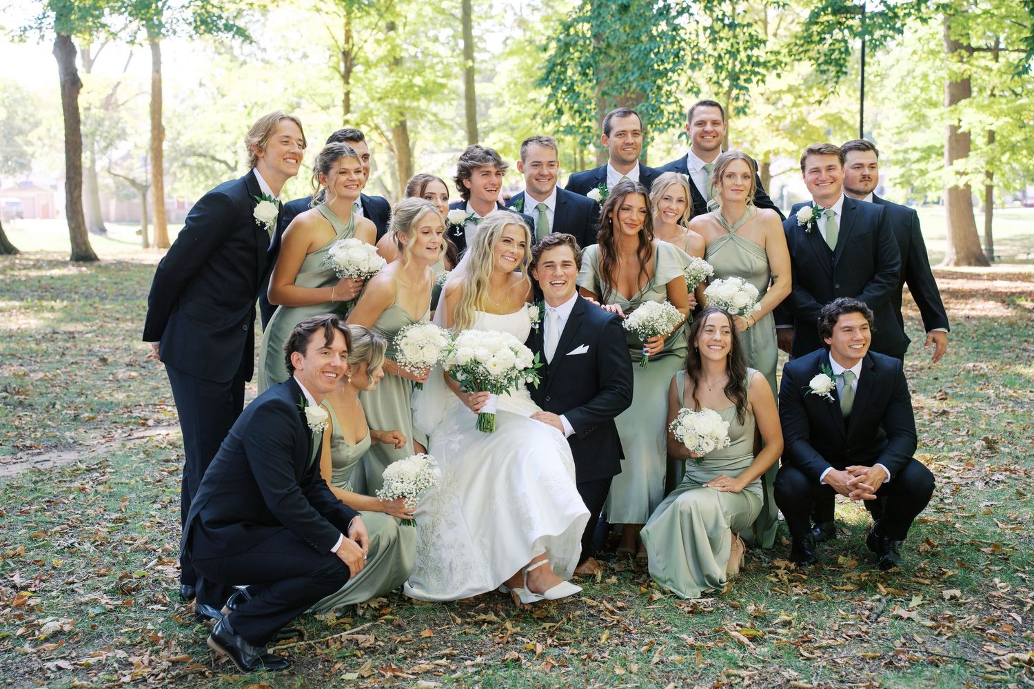 Large wedding party lounges on grass for casual group photo in outdoor setting.