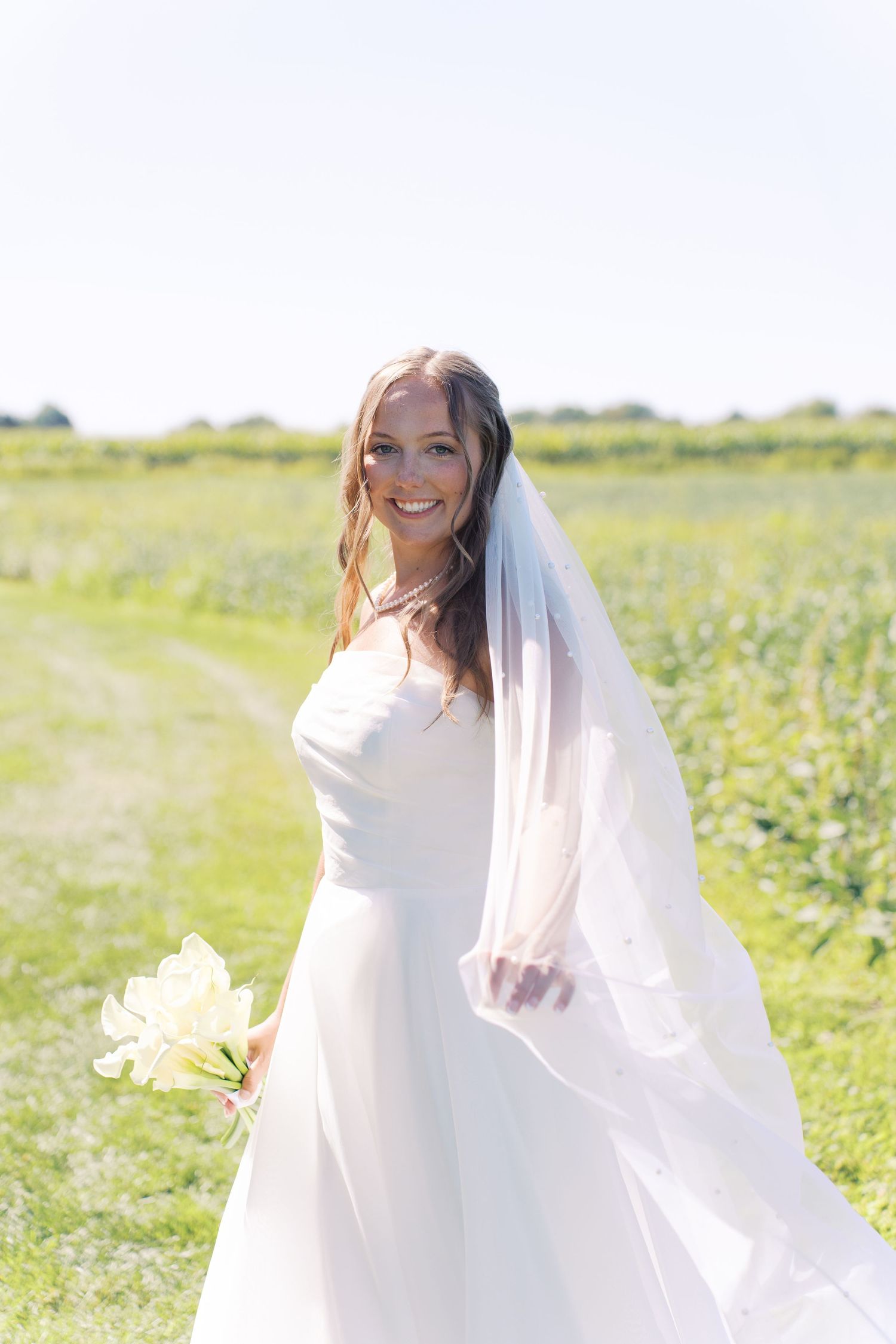 A bride in a white wedding dress poses in a sunny field with her veil flowing.