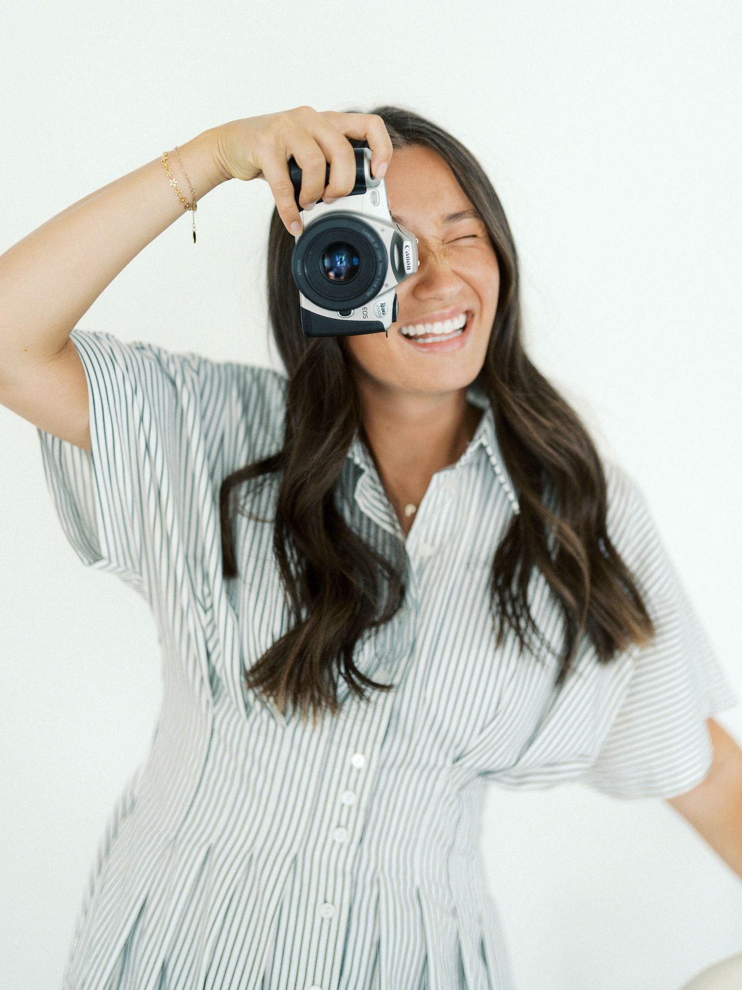 Smiling woman in striped dress holding a camera up to her eye against a white background.