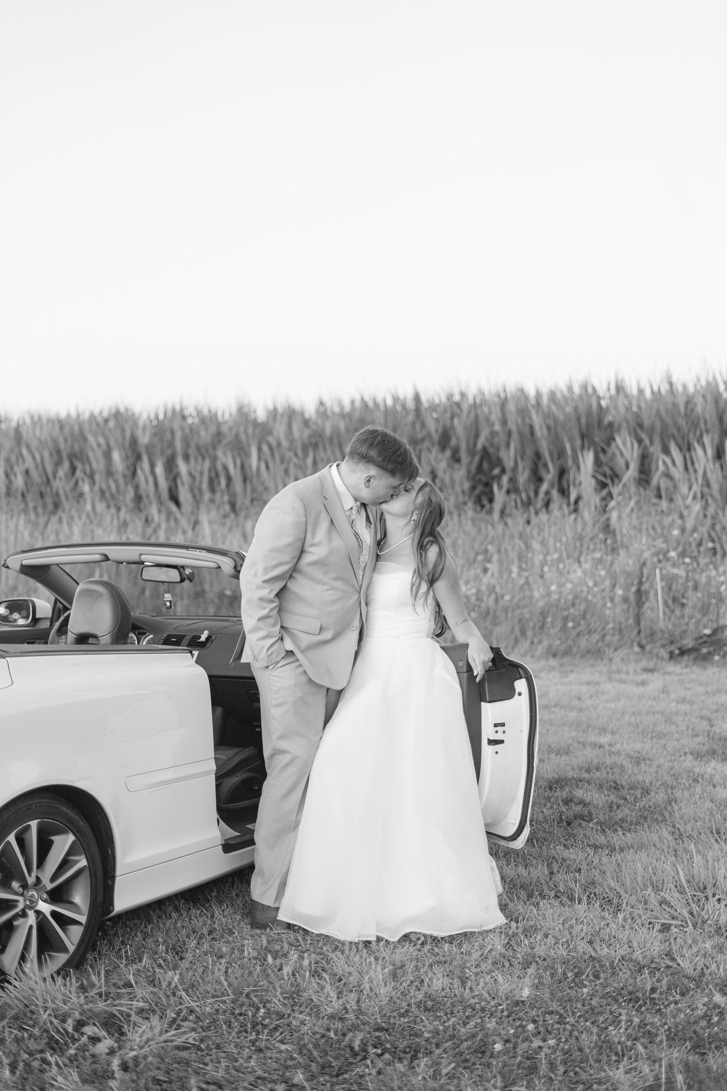 Black and white photo of newlyweds posing with their white car in a cornfield.