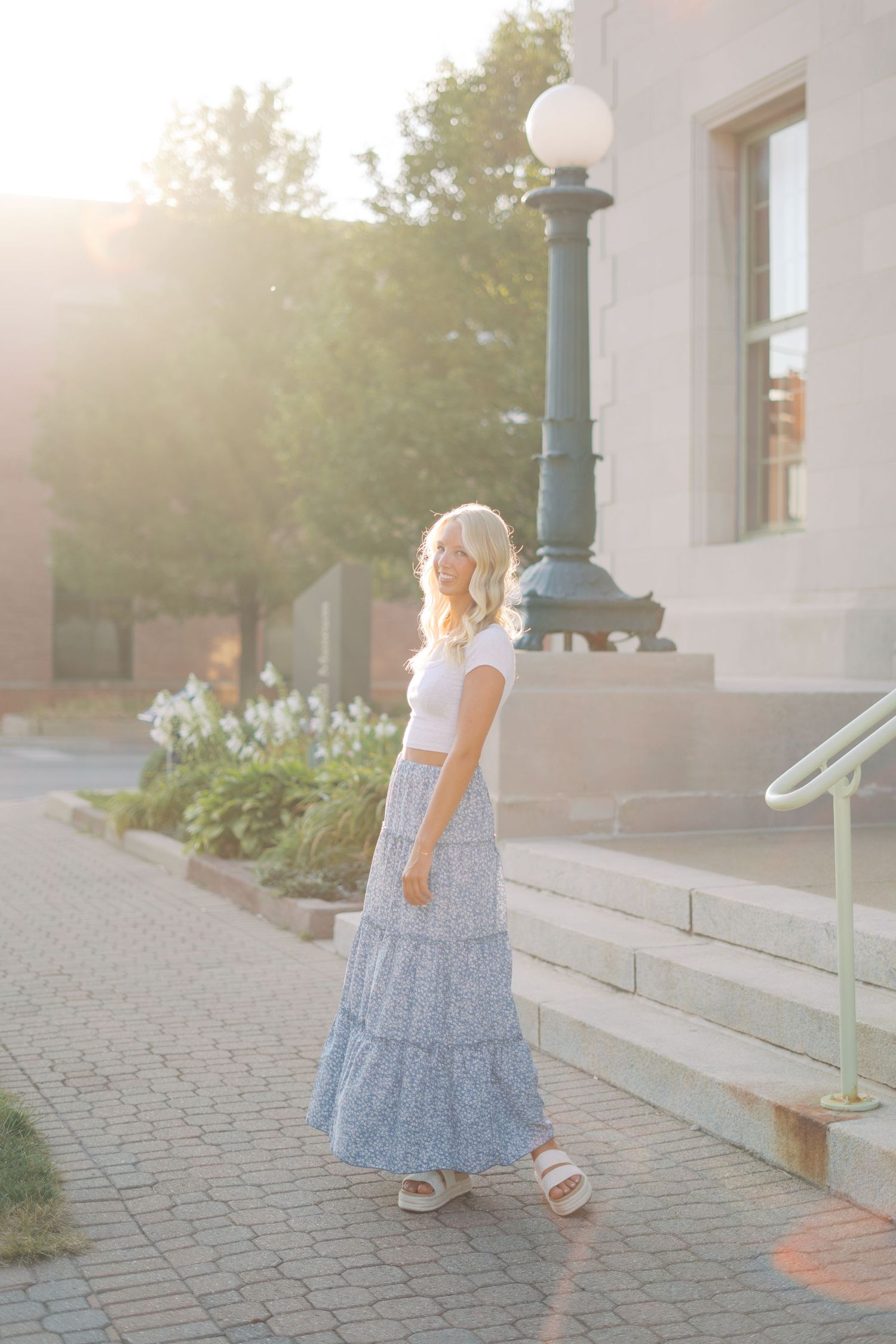 Person in a flowing blue dress walking along a sunlit pathway with architectural elements.