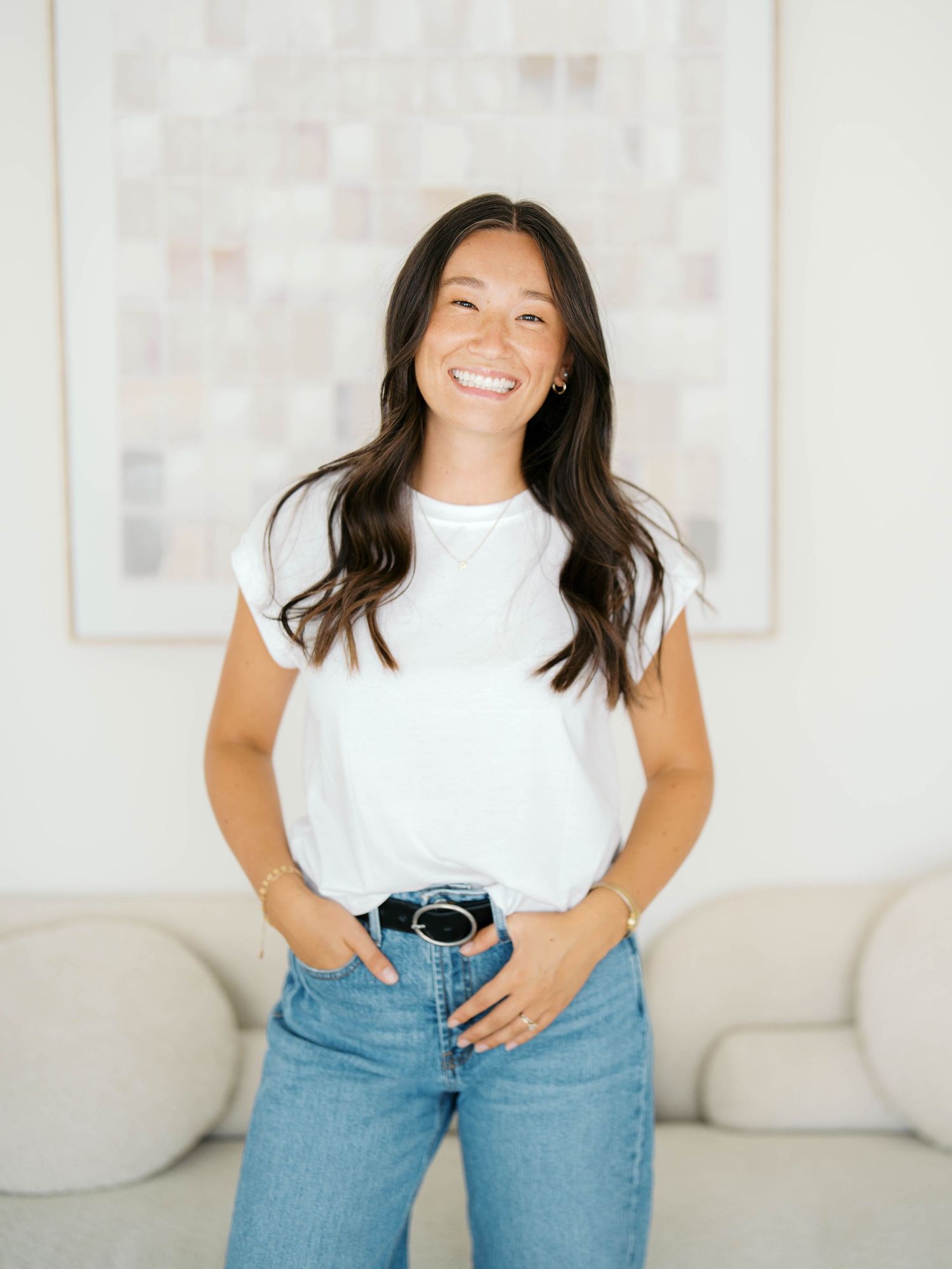 Smiling young woman in white t-shirt and blue jeans standing in a bright modern living room.