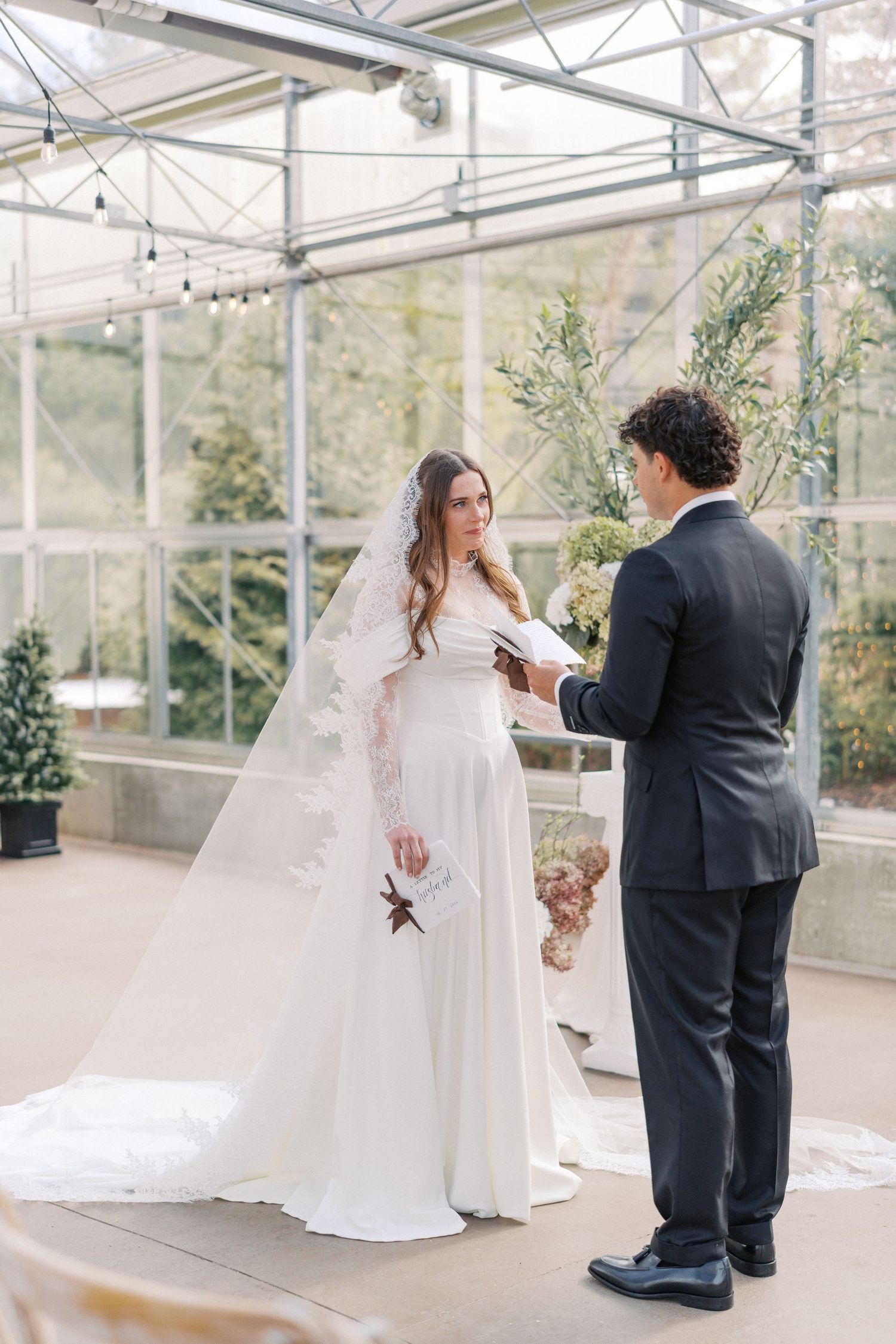 Bride and groom exchange vows in a sunlit greenhouse wedding ceremony, bride wearing a lace veil and white gown.