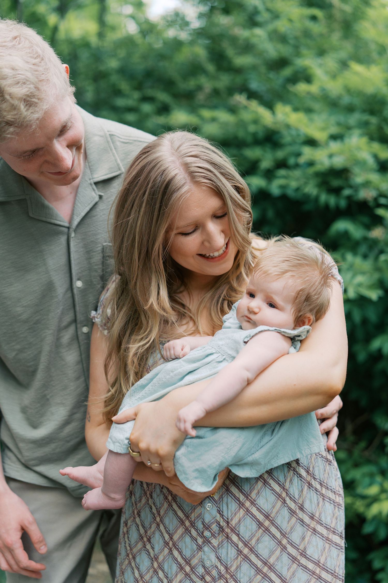 A young family smiles lovingly at their newborn baby outdoors, surrounded by lush green foliage.