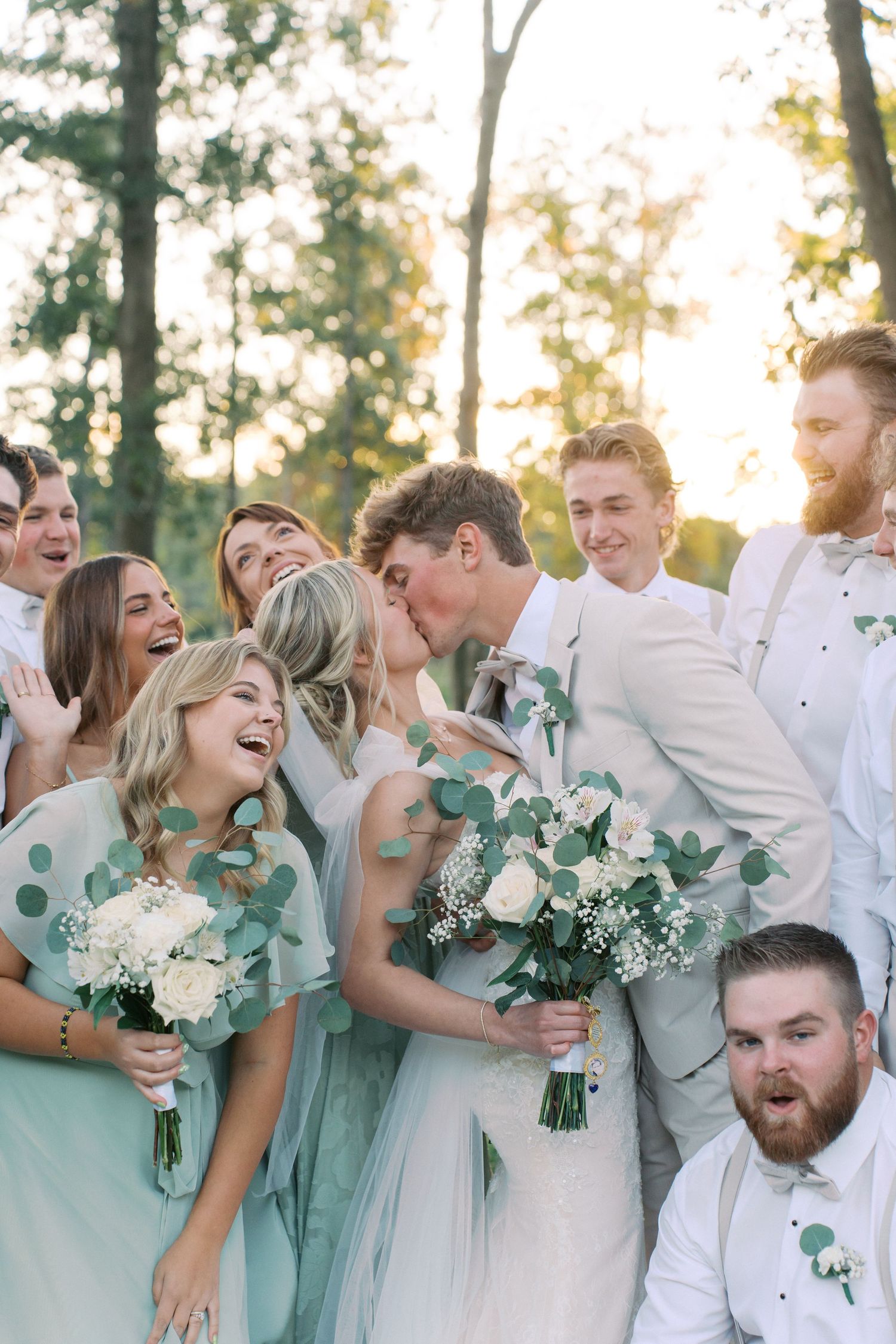 Wedding party in sage green dresses and white suits laughing together outdoors.