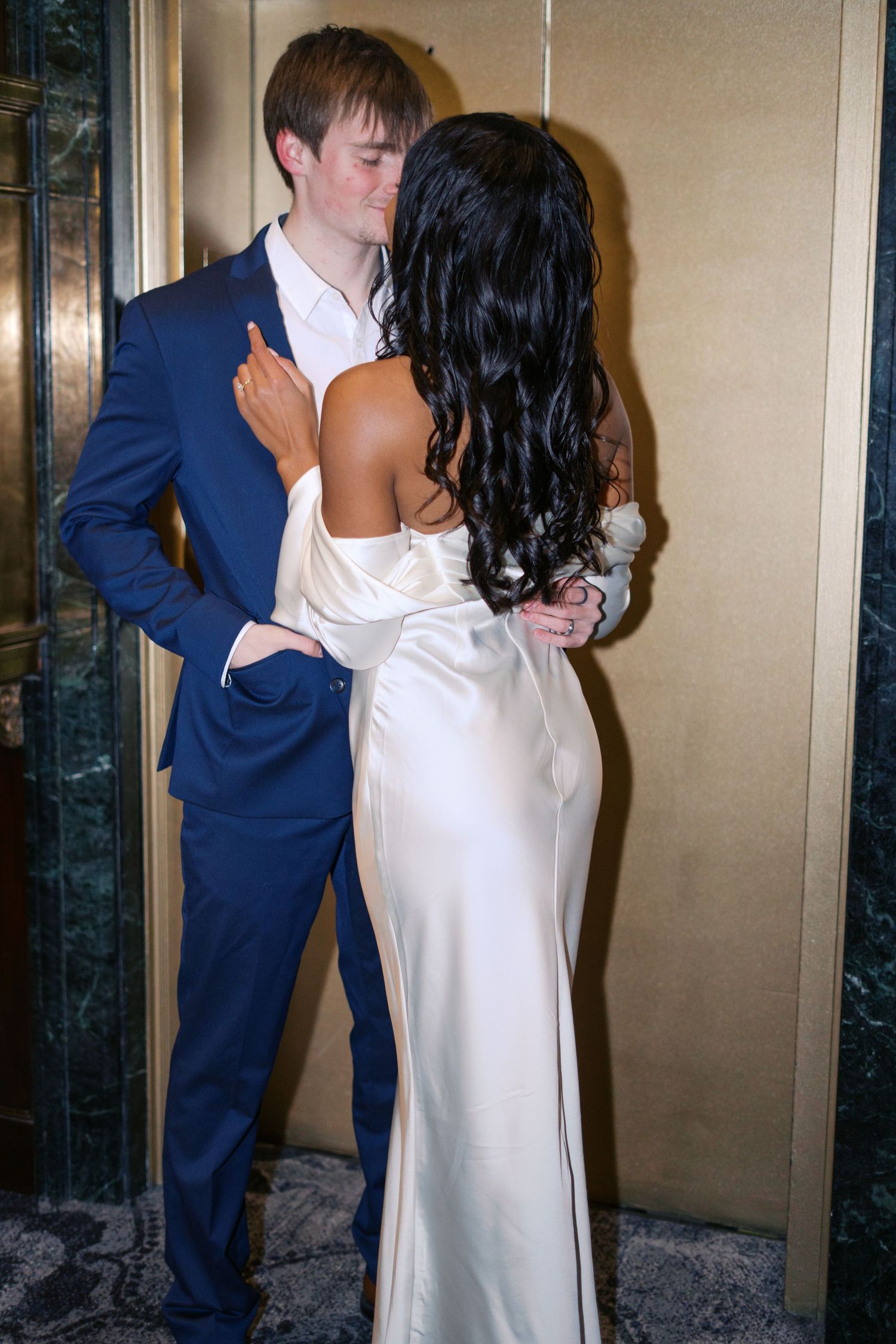 A couple embraces in an elegant indoor setting with gold-toned walls.