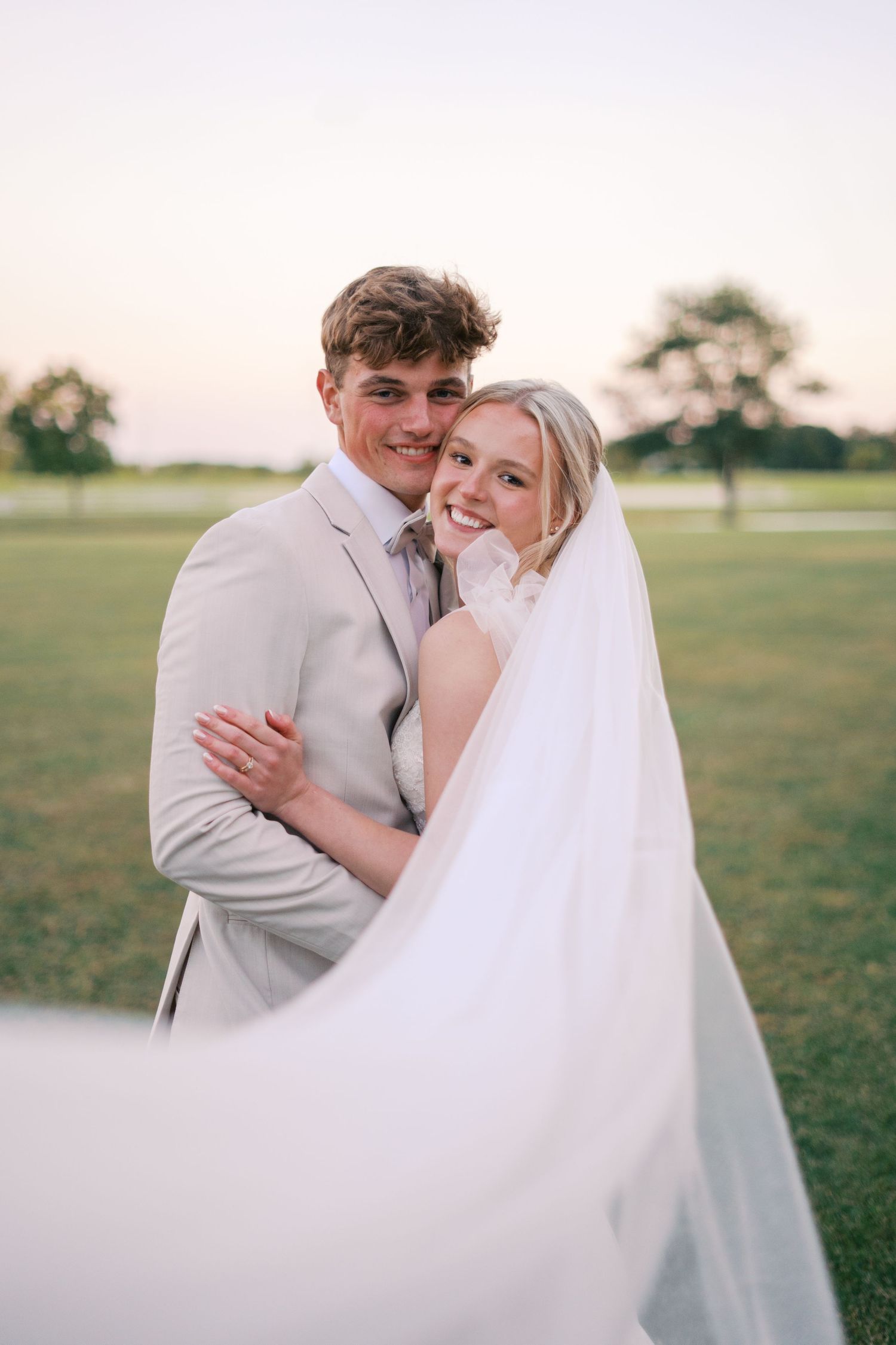 Joyful wedding portrait with flowing veil against sunset field backdrop.