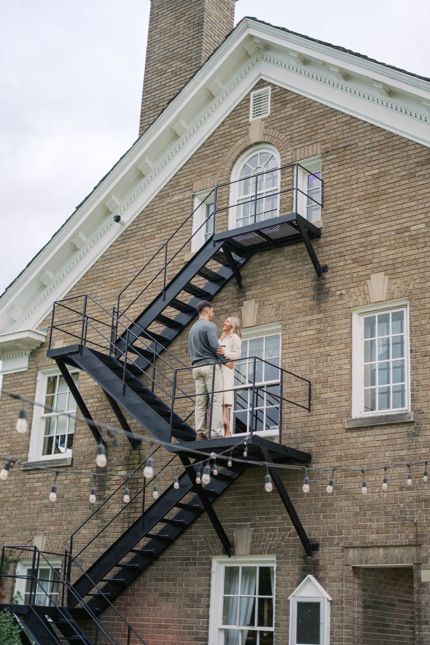 A couple sharing a moment on a black metal fire escape staircase of a brick building.