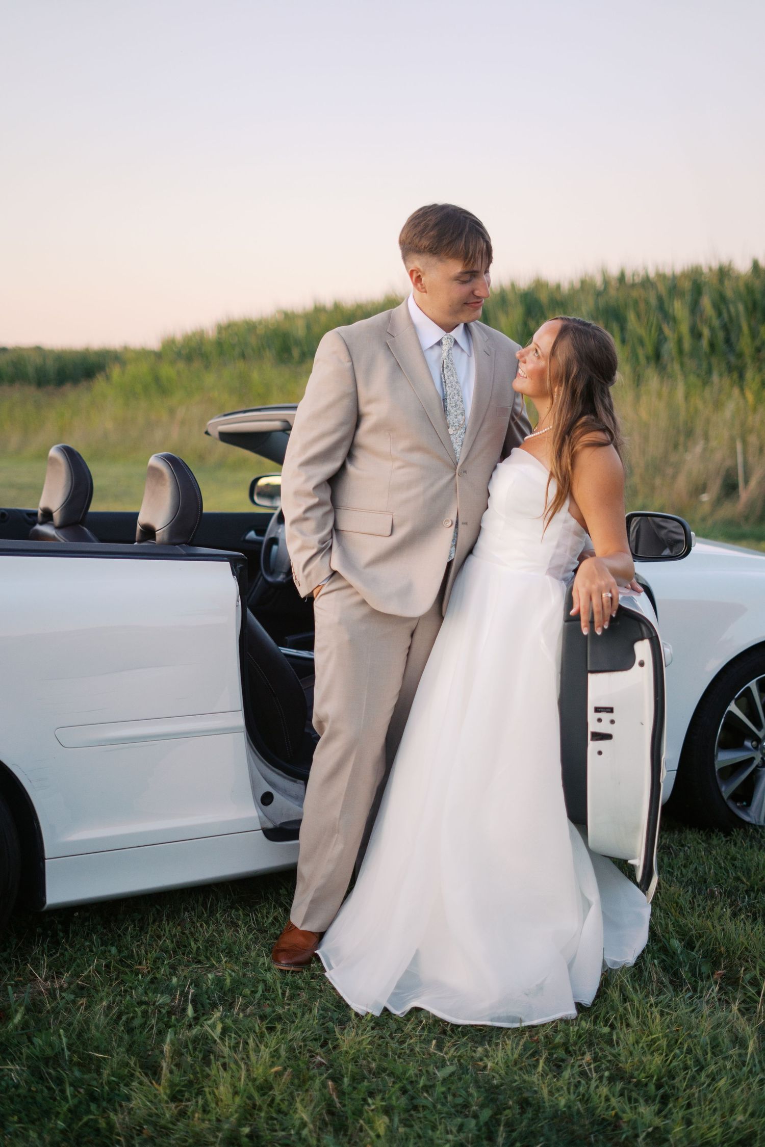 Newlyweds pose playfully against their white car in a rural setting with cornfields.