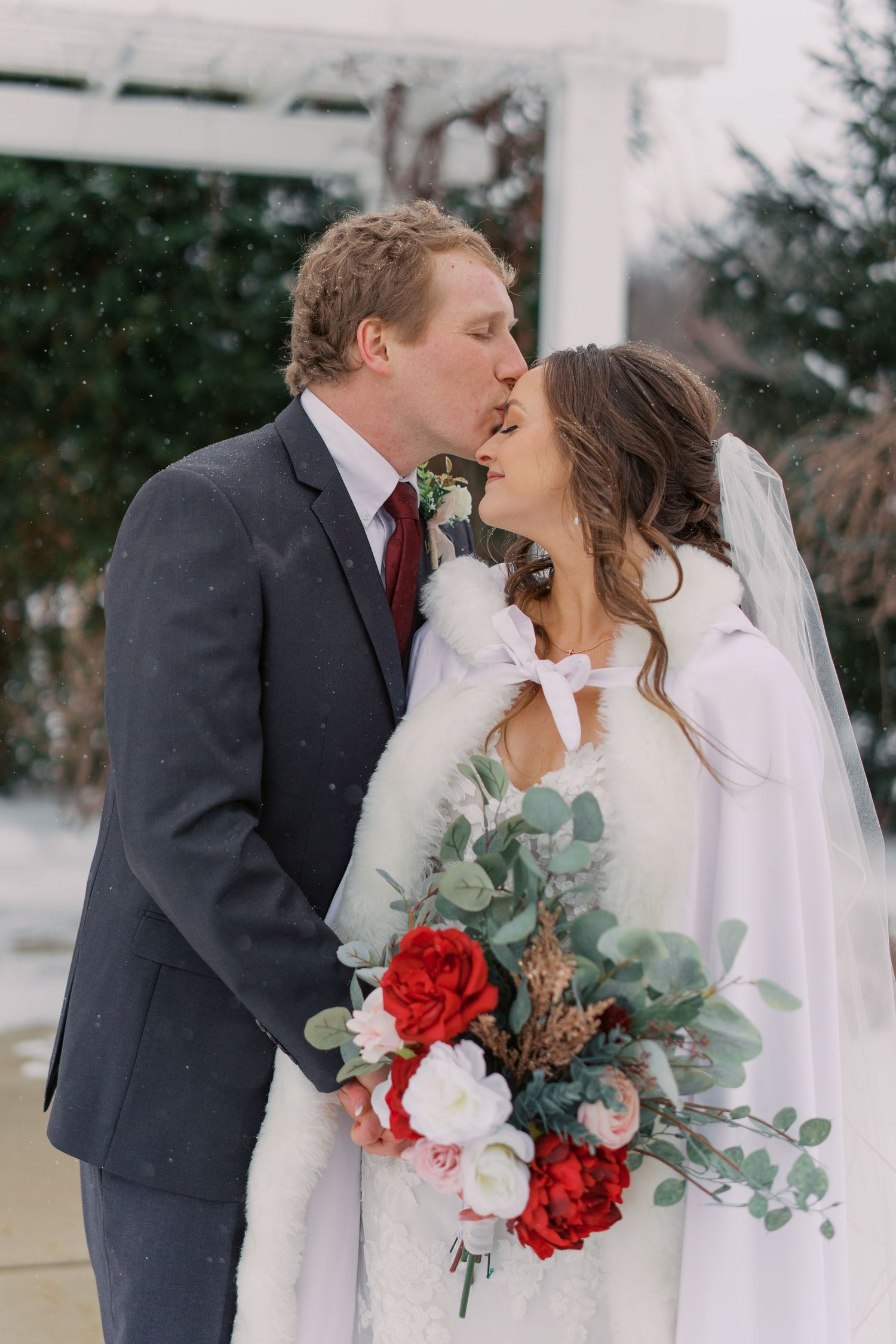 Winter wedding couple kissing while holding red and white bouquet with eucalyptus.