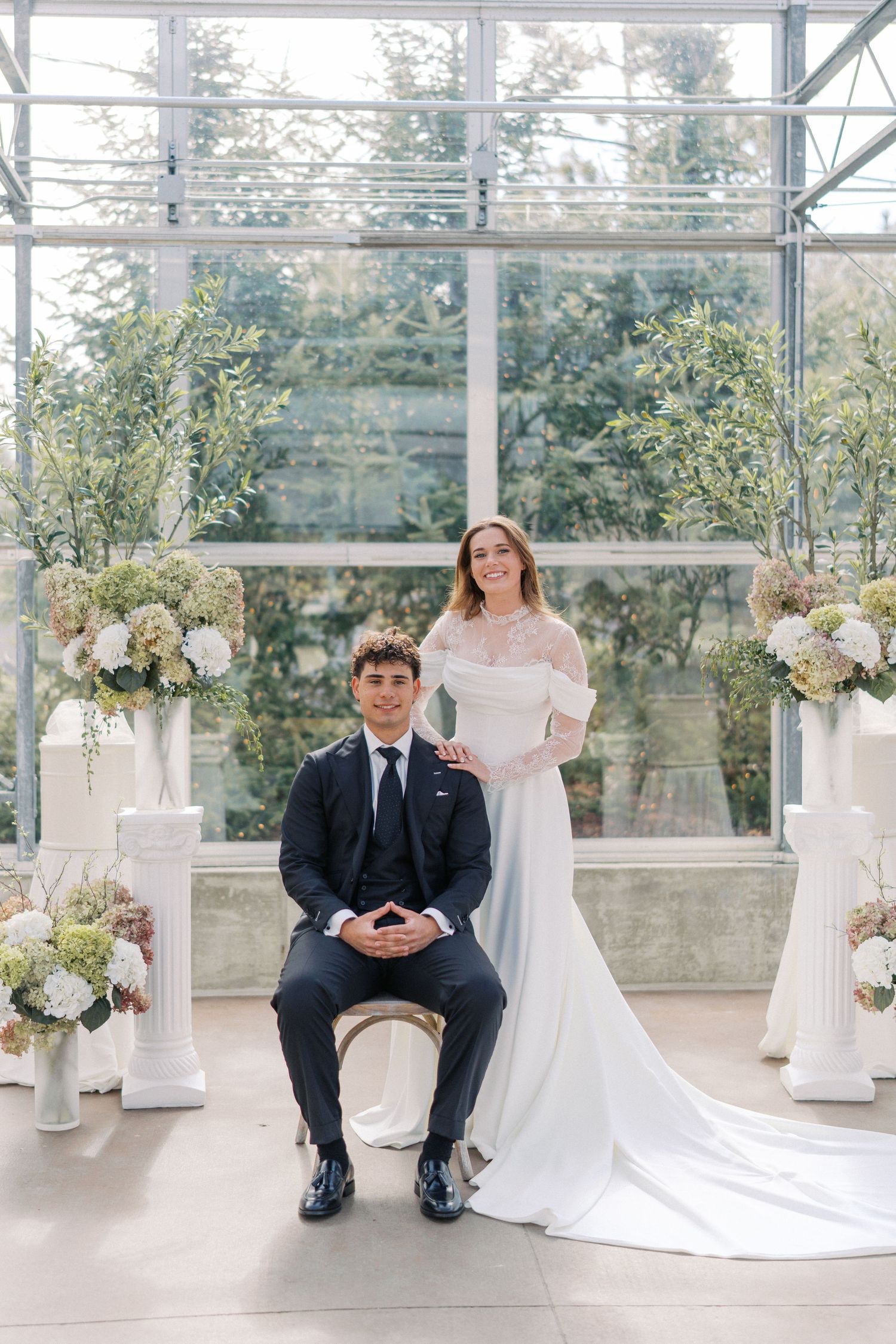 Bride in off-shoulder gown stands behind groom in navy suit seated in elegant greenhouse wedding venue.