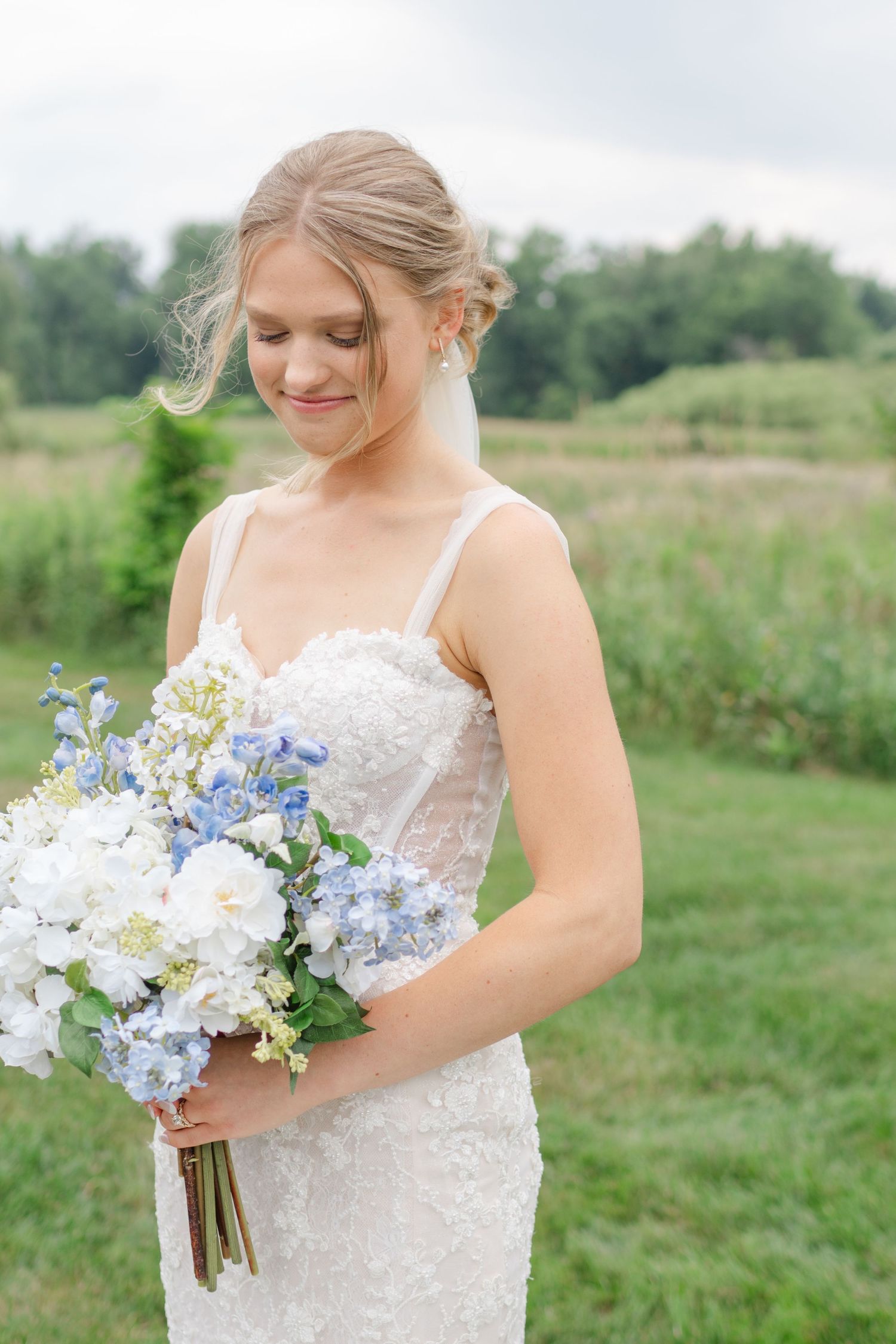 Bride in lace dress holds a beautiful bouquet of blue and white flowers against a pastoral backdrop.