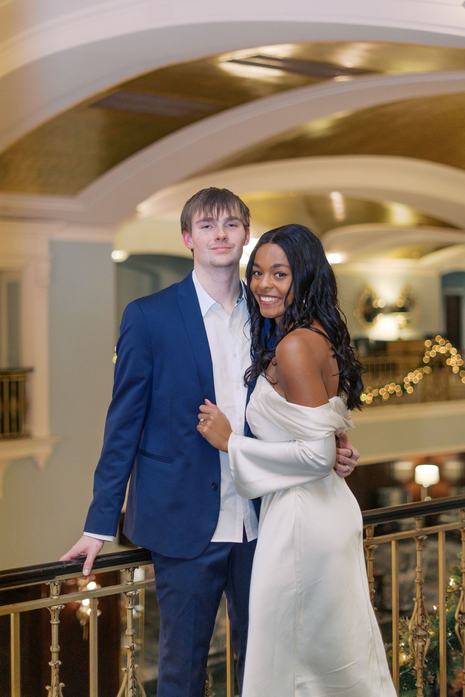 Elegant couple embracing in formal attire near decorated Christmas venue.