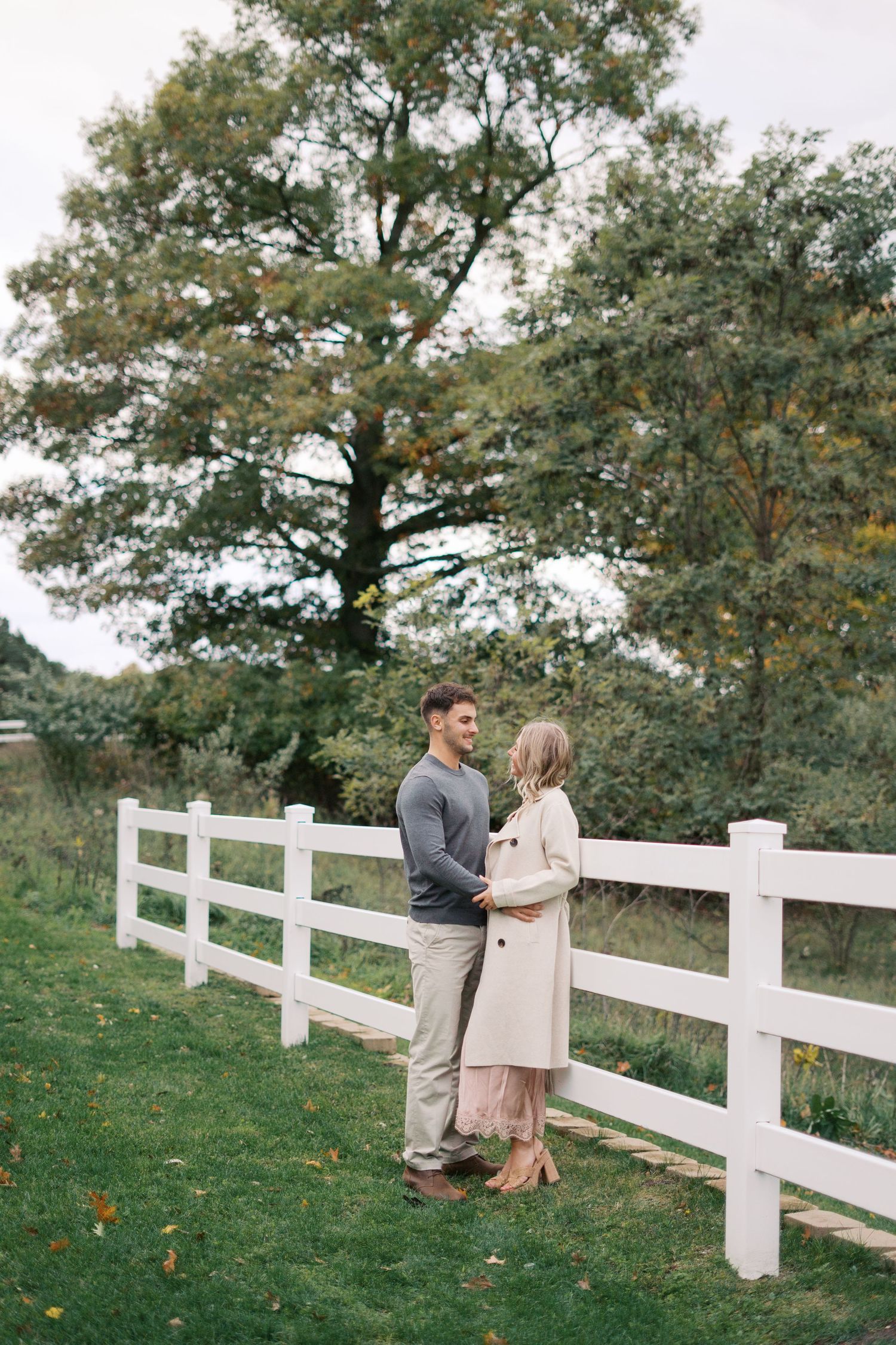 A couple shares a romantic moment by a white fence on a grassy hill during autumn.