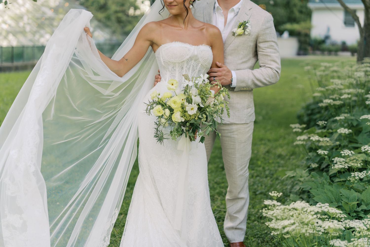 Bride in strapless white gown with flowing veil holds wildflower bouquet beside groom in beige suit outdoors.